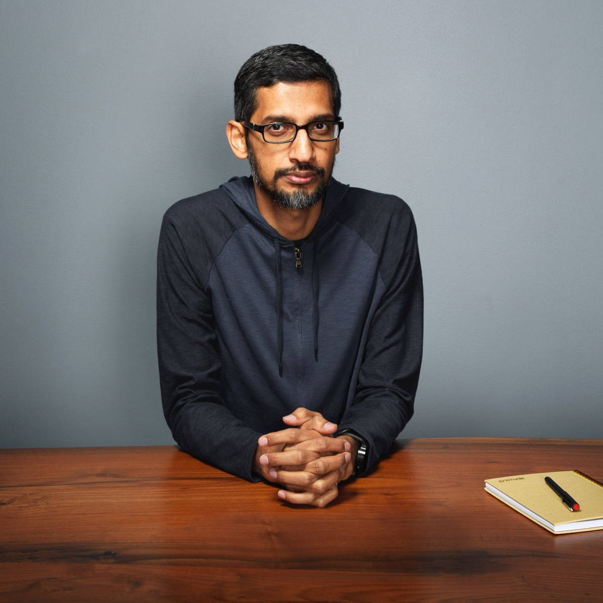 google 桌布 壁紙 — 一個戴眼鏡和鬍鬚的男人坐在木桌旁，穿著深色連帽衫，背景是純灰色牆壁。