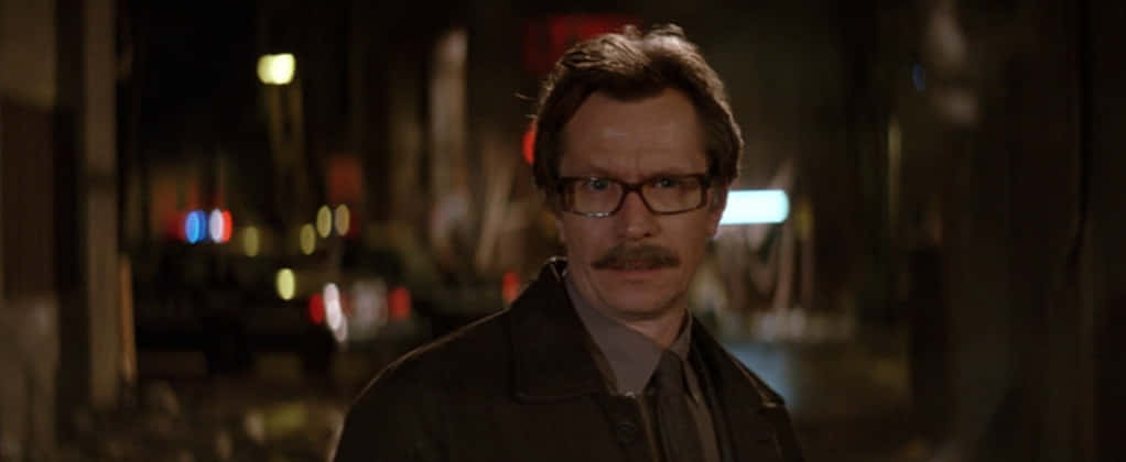 Commissioner Gordon 桌布 壁紙 — 一個戴眼鏡和鬍鬚的男人夜晚站在昏暗的城市街道上，背景有模糊的霓虹燈。