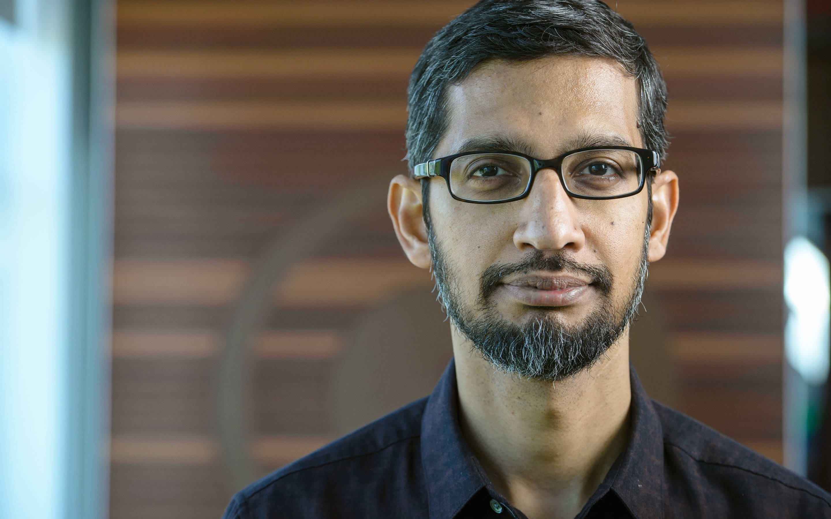 google 桌布 壁紙 — 一個戴眼鏡和鬍鬚的男人穿著深色襯衫的 close-up 肖像，背景是模糊的木質背景。