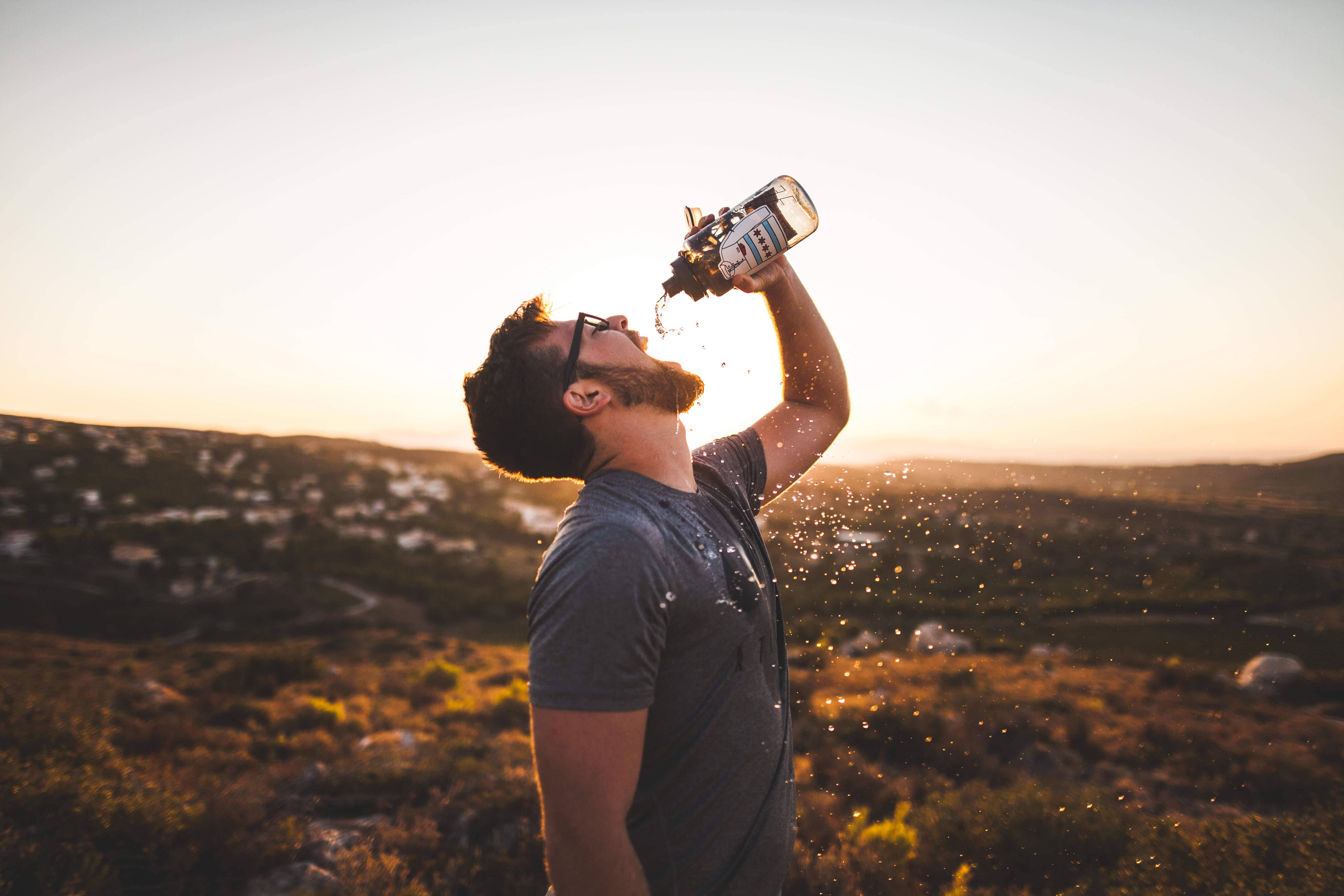 Drinking 桌布 壁紙 — 一個戴眼鏡留鬍鬚的男人，在金色日落下的丘陵景觀中，從瓶子裡喝水。