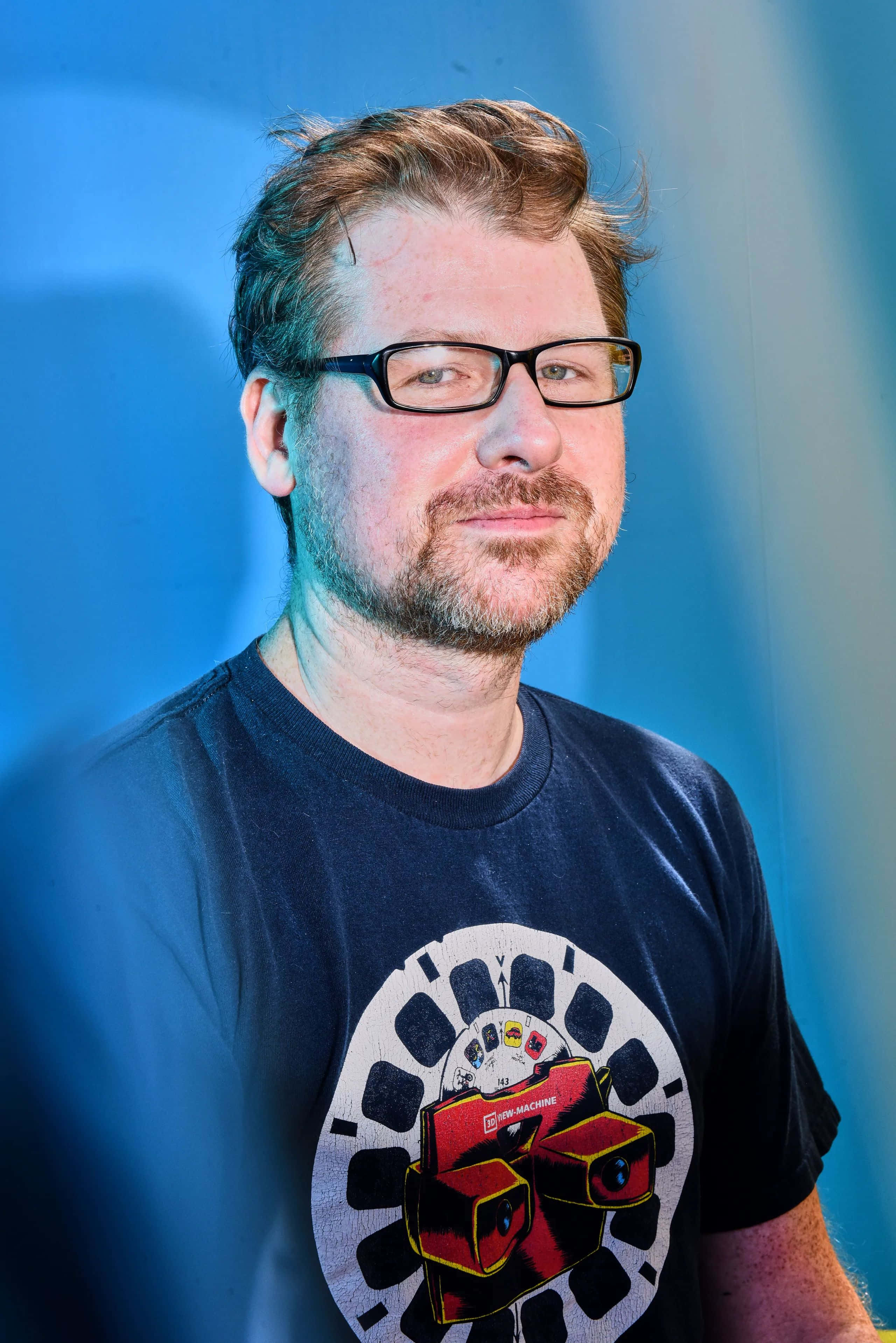 Justin Roiland 桌布 壁紙 — 一個戴眼鏡留鬍鬚的男人，身穿深色T恤，上面有紅白圖案，背景為藍色。