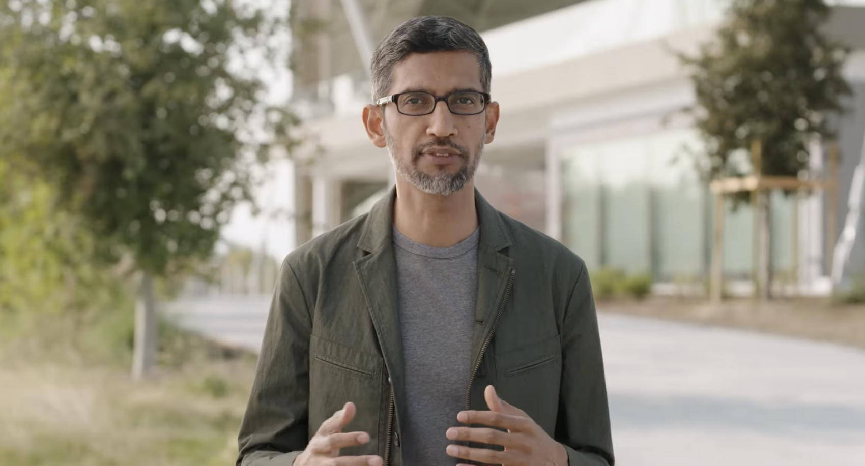 google 桌布 壁紙 — 一個戴眼鏡留鬍鬚的男人站在室外一座現代建築前面，一邊說話一邊做手勢。