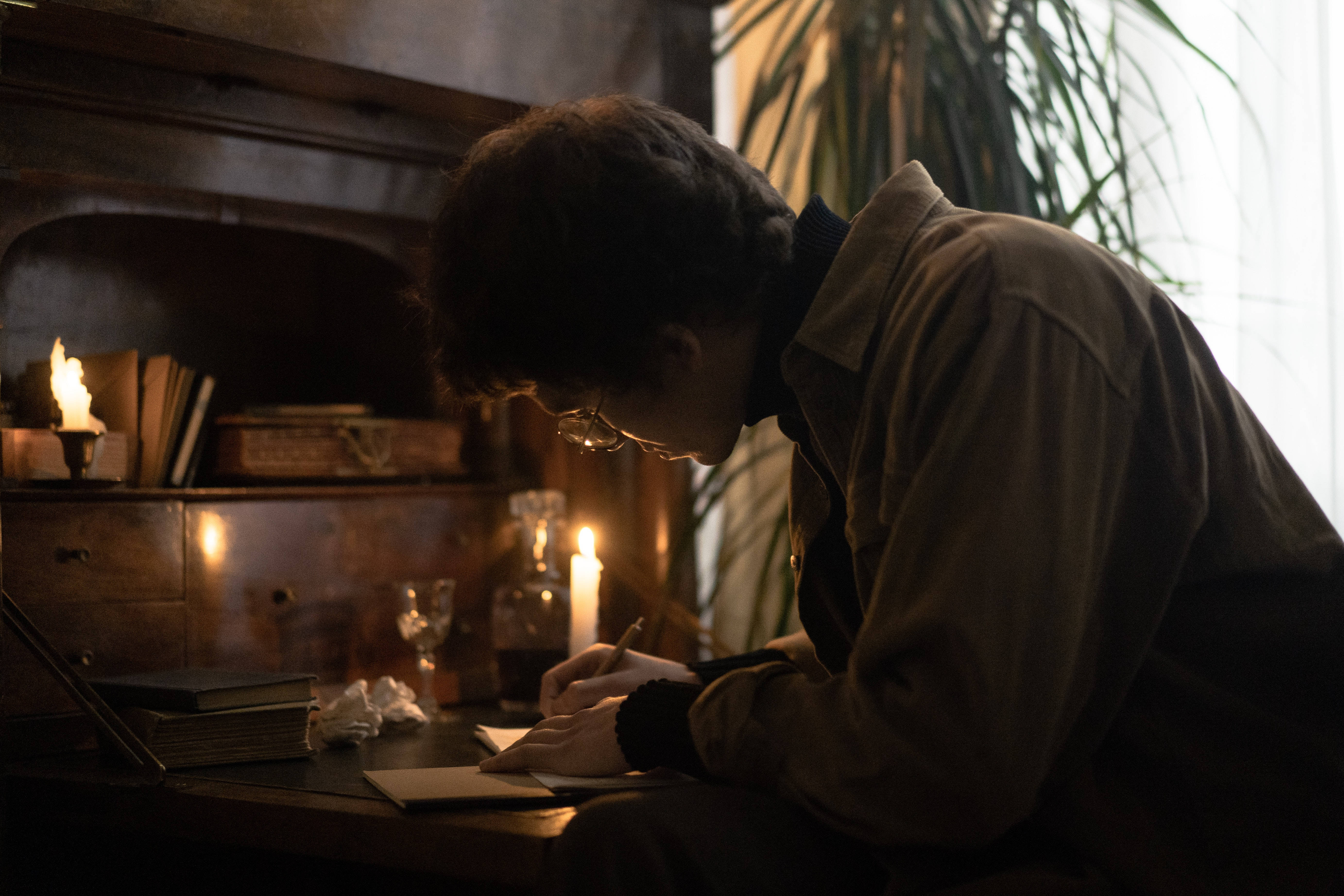 暗黑學院風 桌布 壁紙 — 一個戴眼鏡的人在一間昏暗的復古房間裡，在木桌上用燭光寫字，房間裡有一個壁爐。