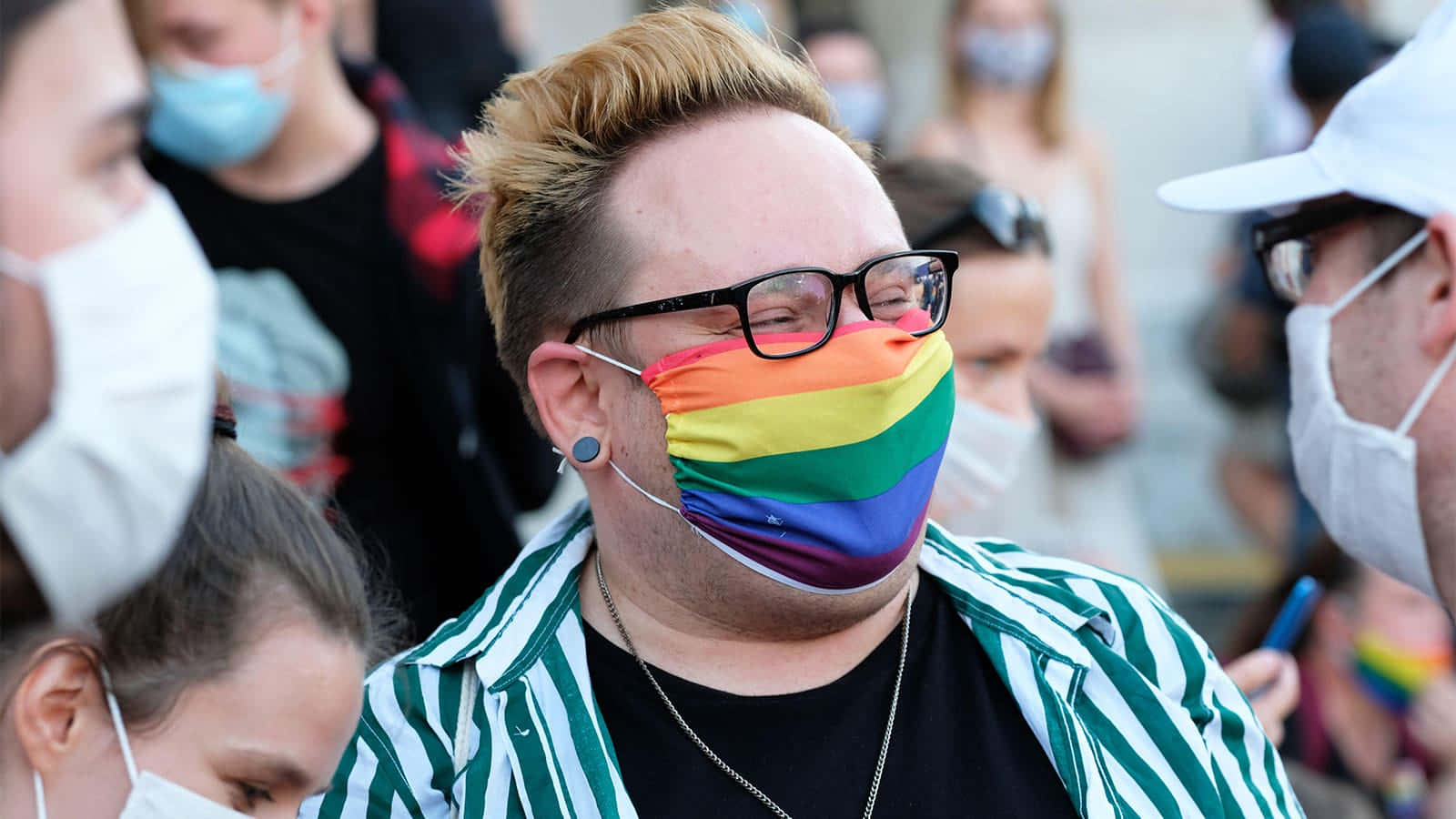 男同志 桌布 壁紙 — 一個戴眼鏡的人在擁擠的戶外Pride活動中戴著彩虹口罩，周圍還有其他戴口罩的人。