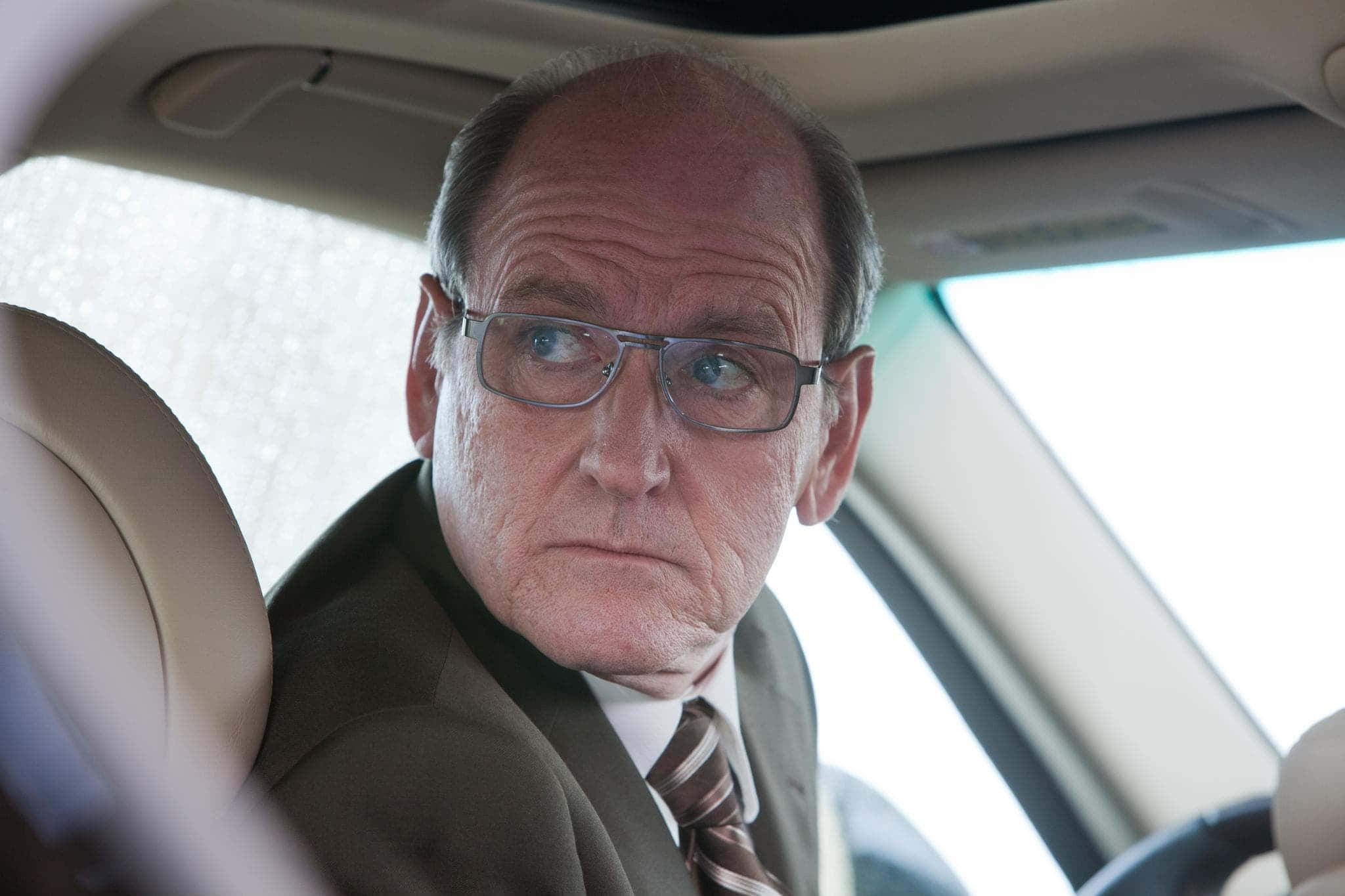 Richard Jenkins 桌布 壁紙 — 一個戴眼鏡的嚴肅中年男子坐在車內，透過雨水模糊的車窗專注地向前看。
