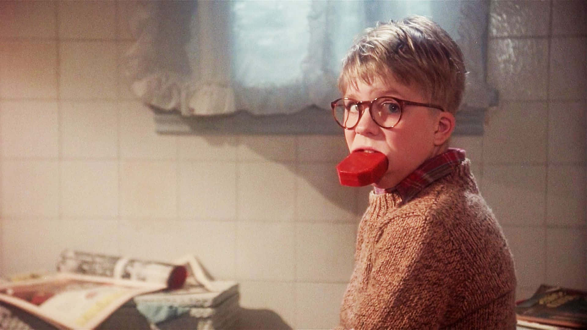 a christmas story 桌布 壁紙 — 一個戴眼鏡的小男孩在陽光充足的廚房裡，拿著紅色電話聽筒貼在嘴邊，牆上鋪了瓷磚，還有報紙。