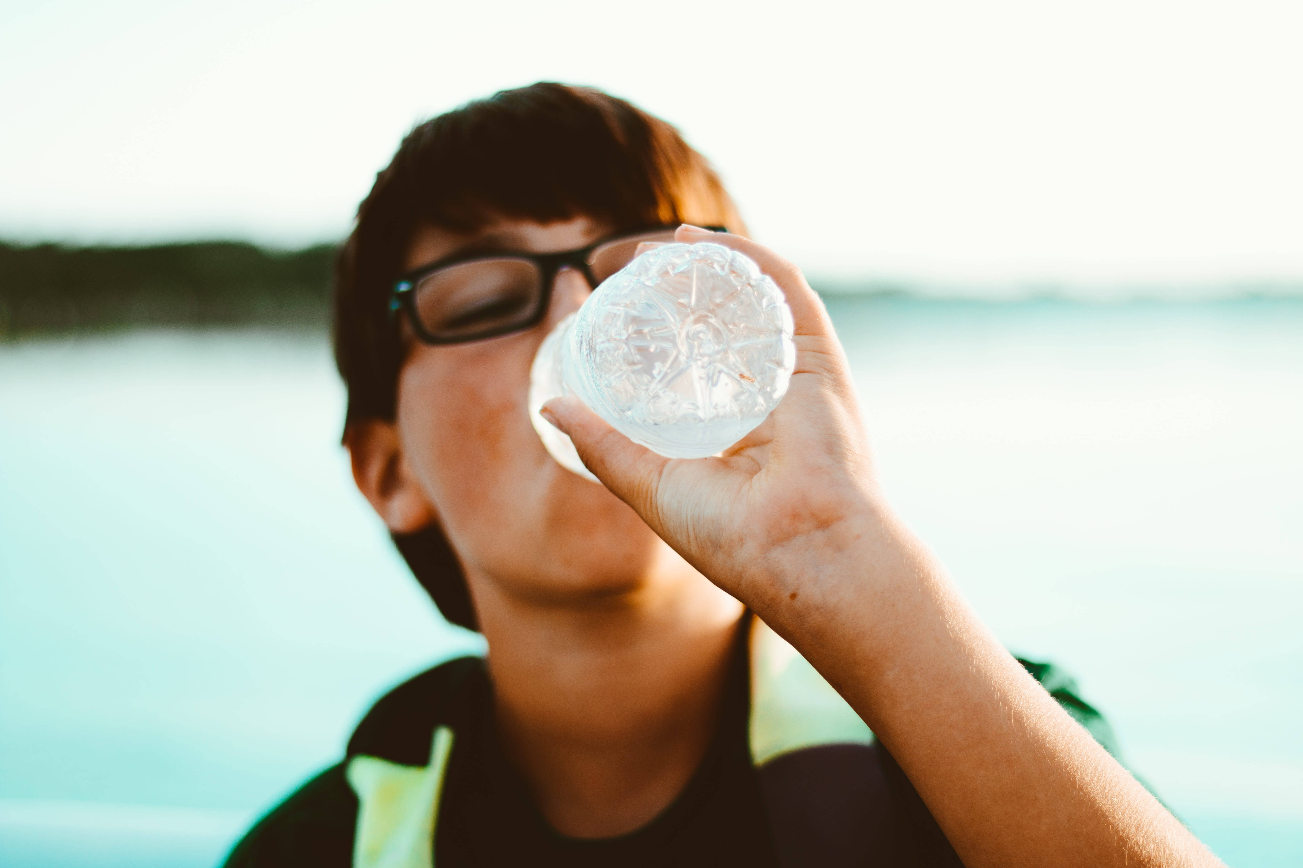 水 桌布 壁紙 — 一個戴眼鏡的年輕人在柔和模糊的湖景背景下，用透明塑膠瓶喝水。