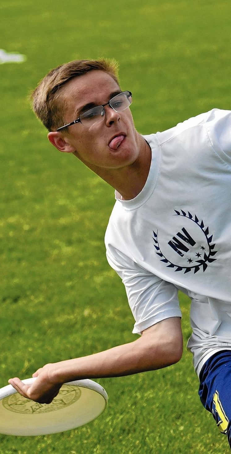 Athlete 桌布 壁紙 — 一個戴眼鏡的年輕男子在充滿活力的綠色場地上抓住飛盤，頑皮地伸出舌頭。