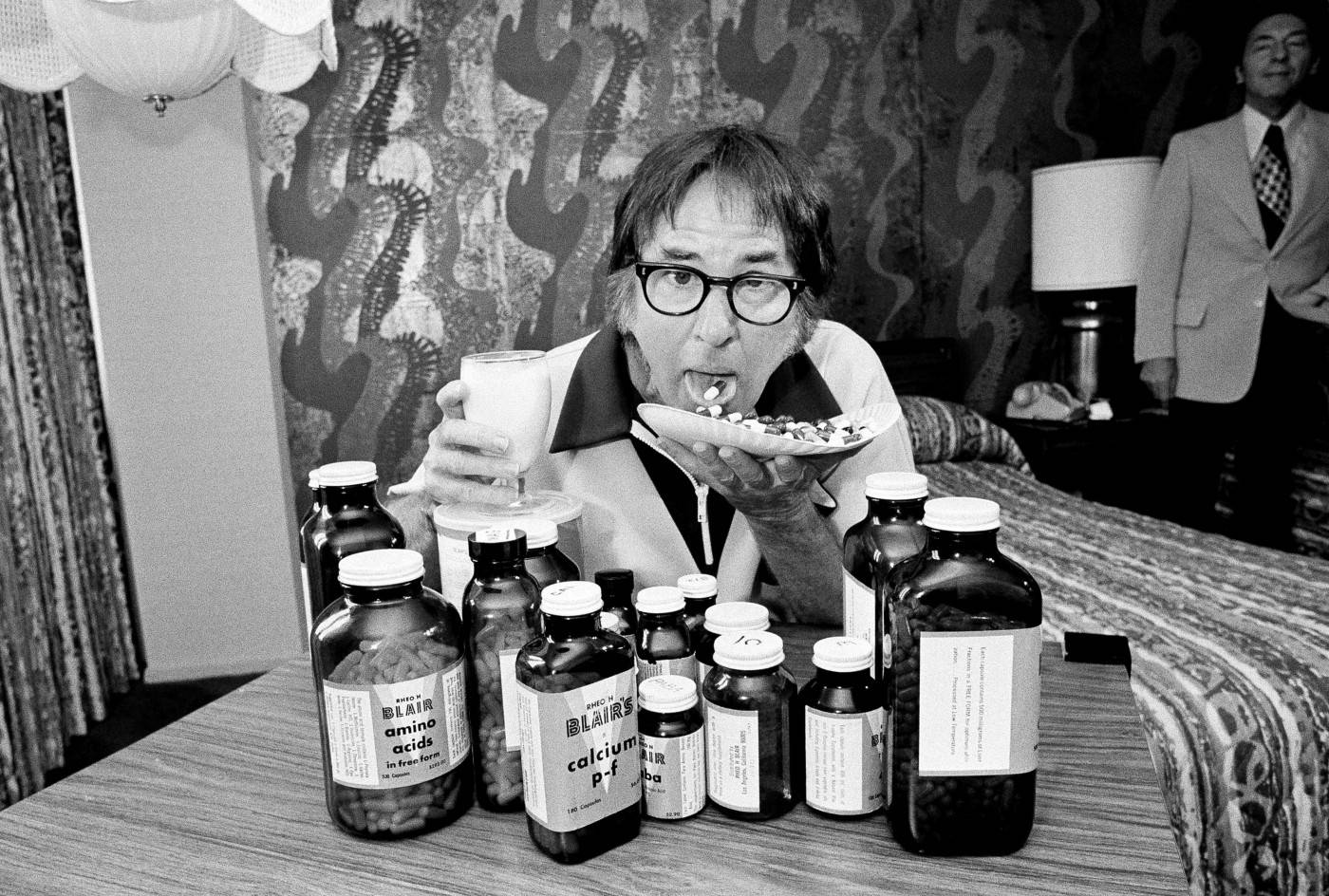 Man 桌布 壁紙 — 一個戴眼鏡的男人在一個有圖案壁紙的房間裡，拿著一杯牛奶和一碟藥丸，周圍是補充劑瓶。