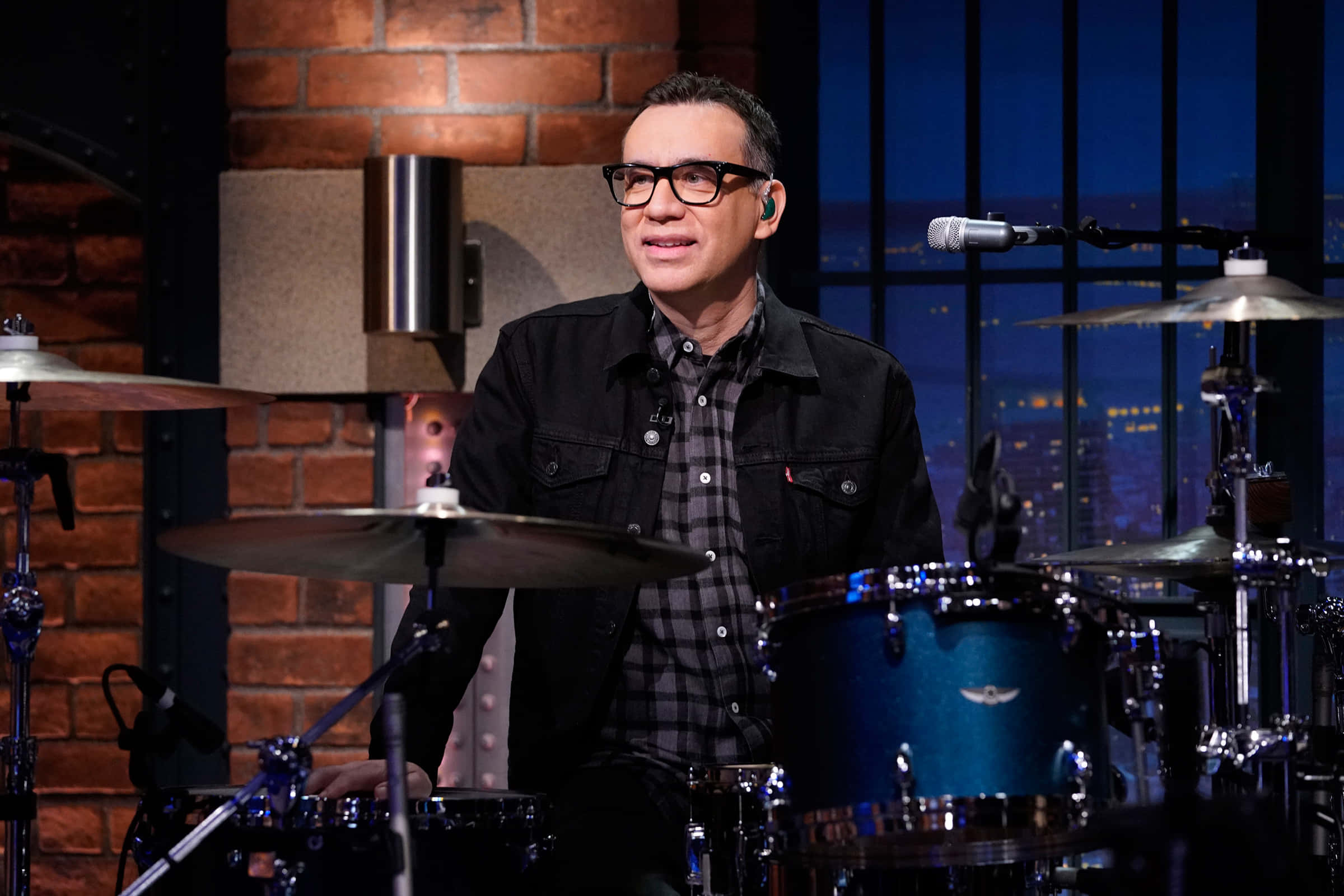 fred armisen 桌布 壁紙 — 一個戴眼鏡的男人坐在一個有磚牆和透過窗戶可見夜間城市天際線的錄音室裡的鼓組後面。