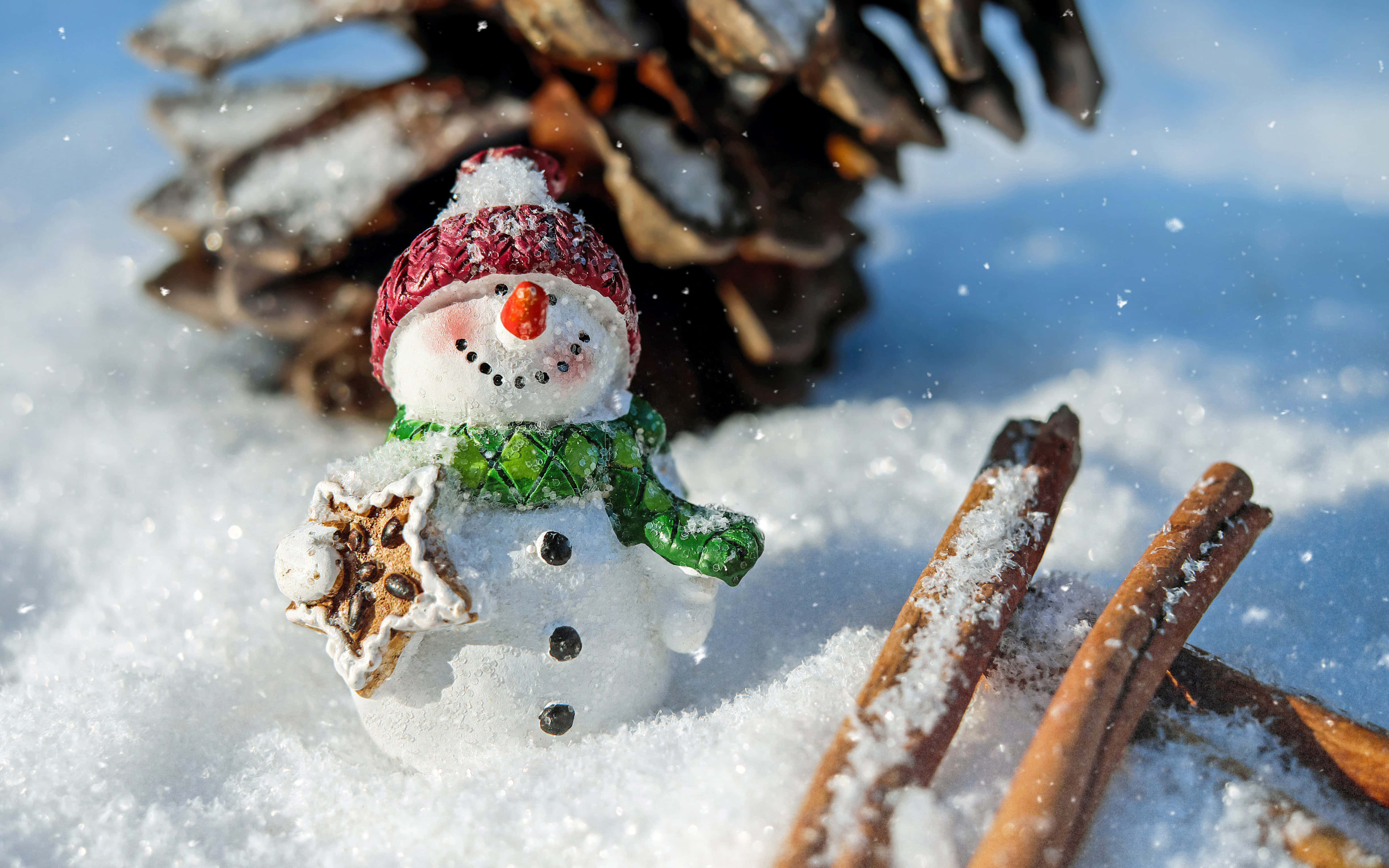 Christmas Snow 桌布 壁紙 — 一個戴紅帽、圍綠圍巾的開心雪人公仔，在新鲜雪地上拿著一個星形餅乾，旁邊有松果和肉桂棒。