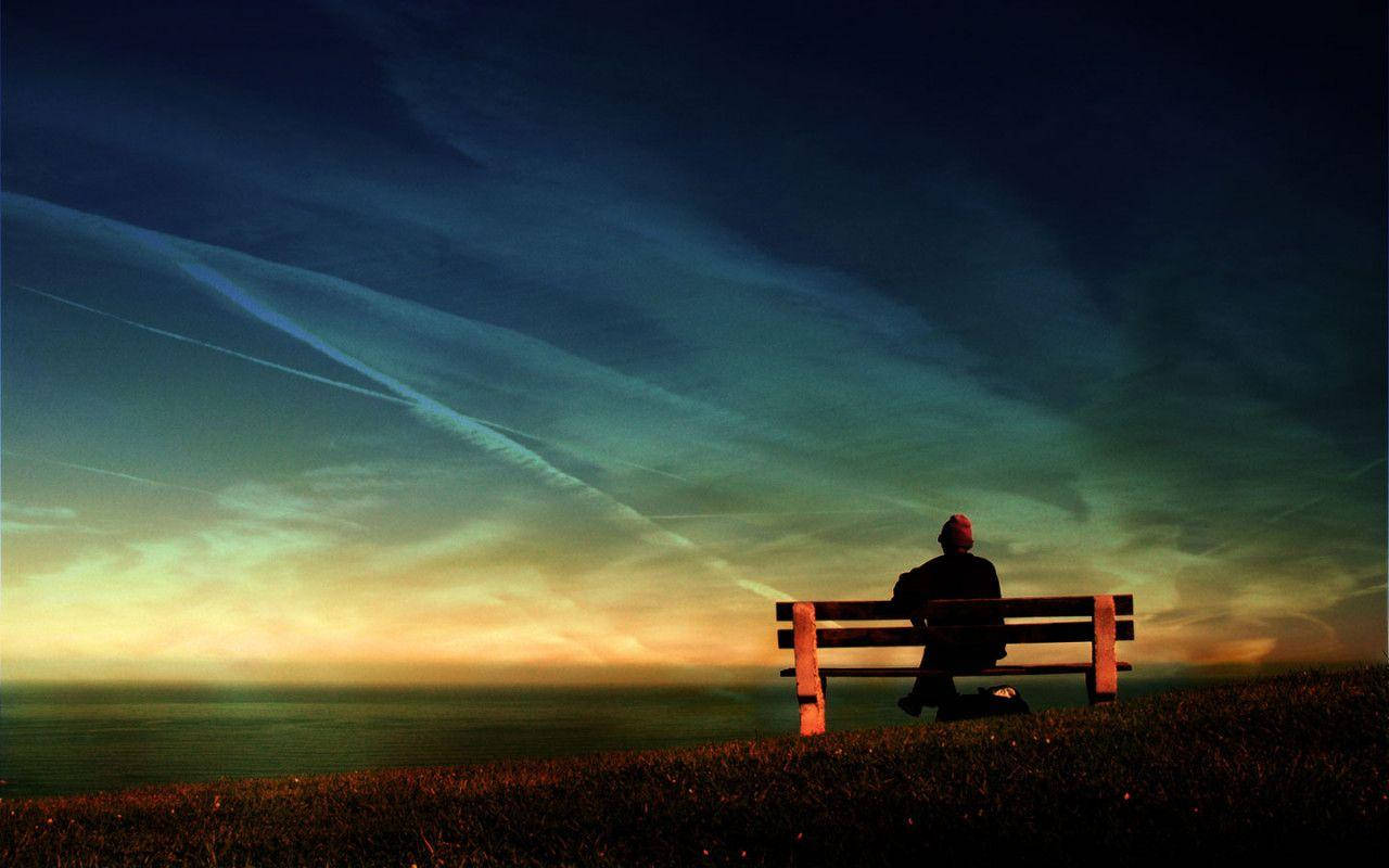 Alone Boy 桌布 壁紙 — 一個戴紅帽的孤獨身影坐在木長椅上，俯瞰暮色中廣闊的海洋，天空有條紋雲和溫暖的金色地平線。