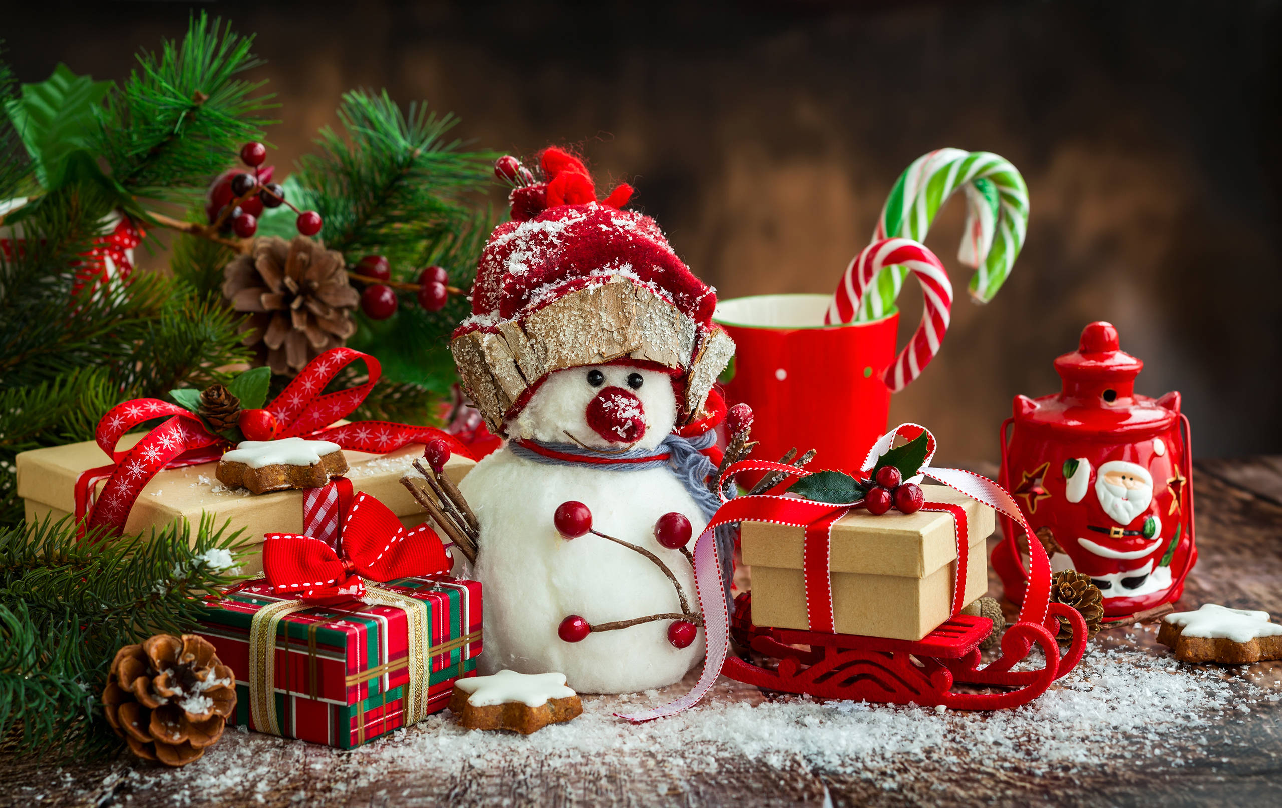 Presents 桌布 壁紙 — 一個戴紅帽的節日雪人公仔，旁邊放著包裝好的禮物、拐杖糖和松樹枝，放在鋪滿雪的木桌上。