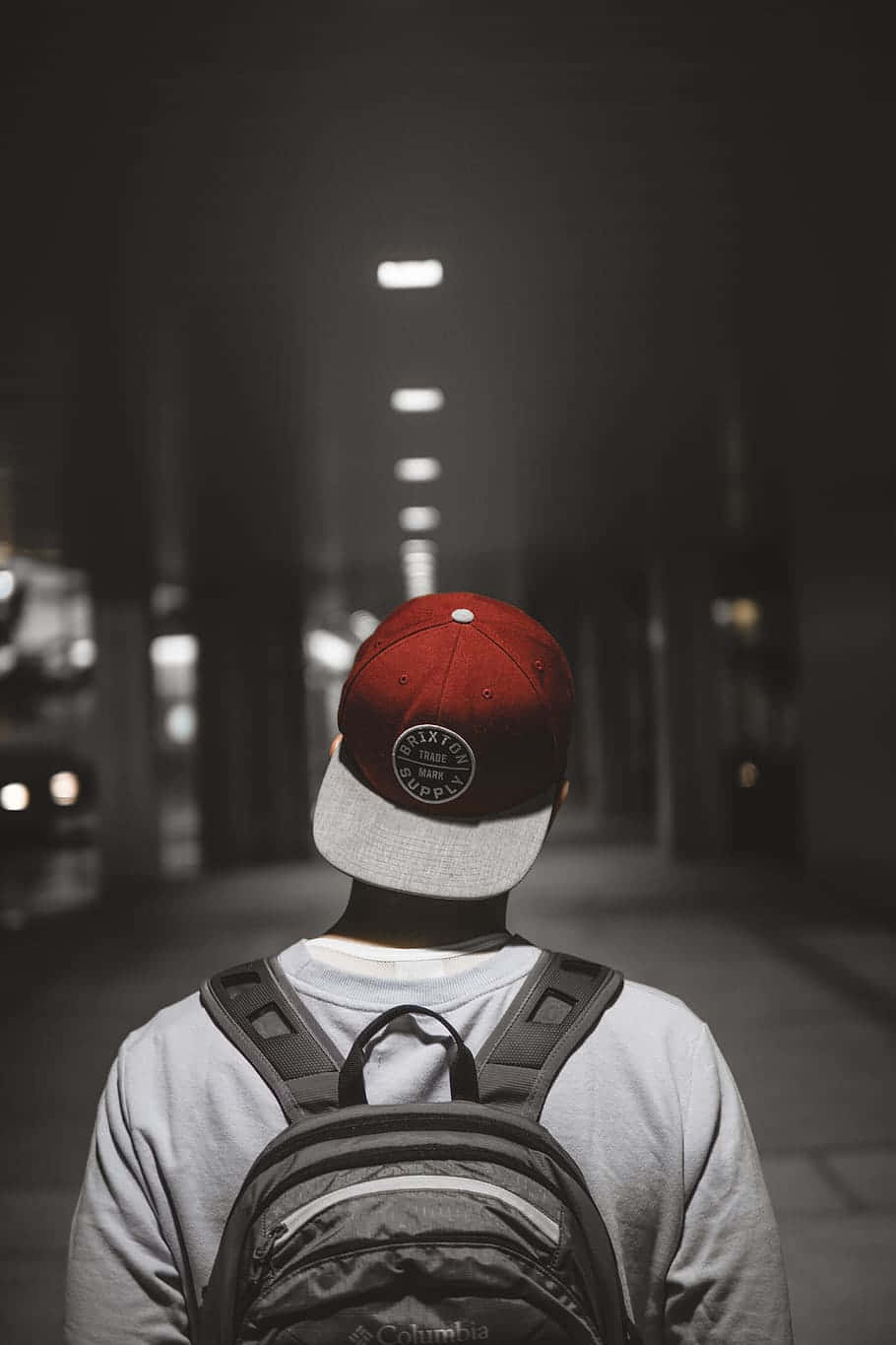 攝影 桌布 壁紙 — 一個戴紅色帽子和背包的人，在夜晚沿著燈光昏暗的城市街道行走，前方有發光的燈光逐漸遠去。