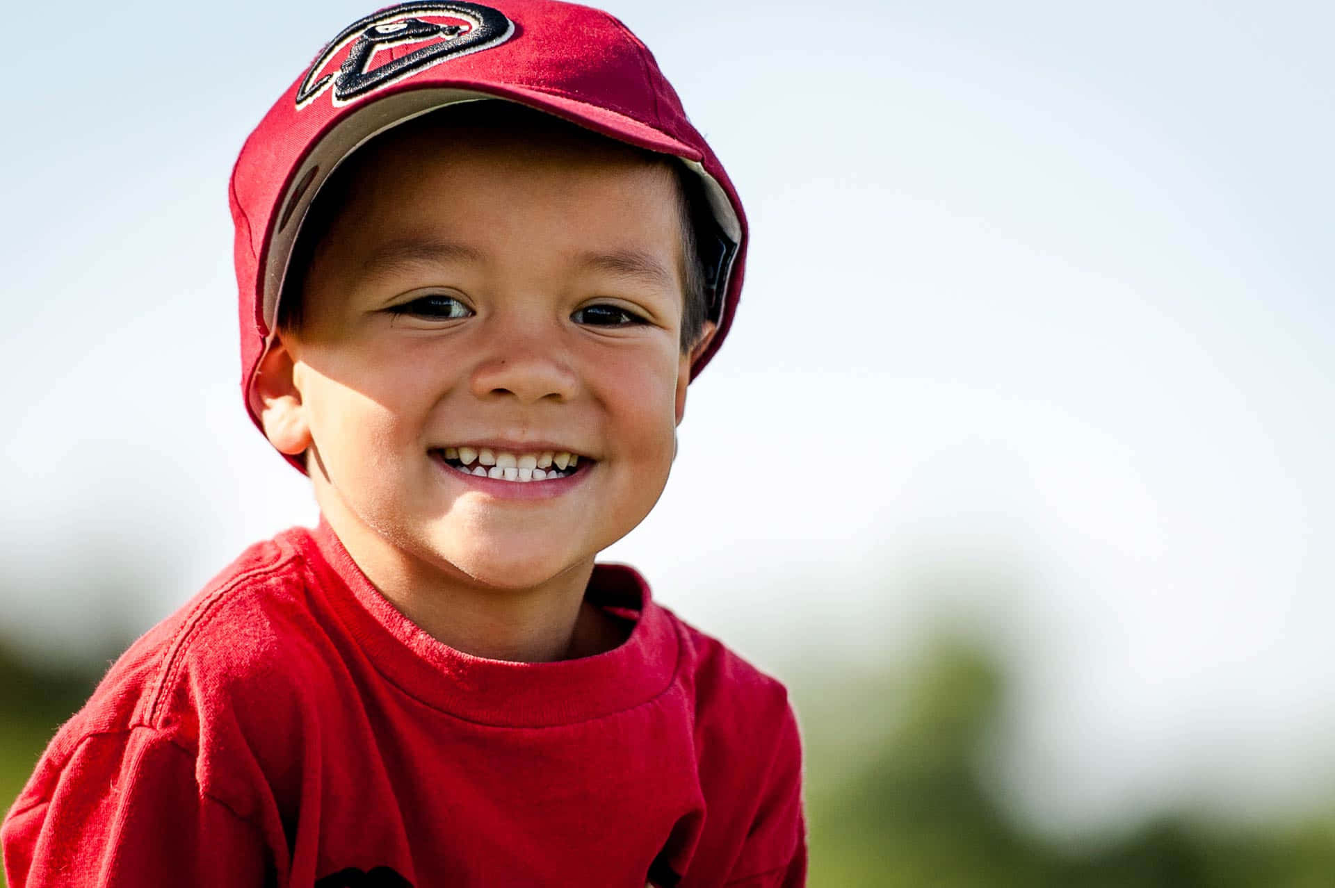 Little League Baseball 桌布 壁紙 — 一個戴紅色棒球帽、穿紅色襯衫的開朗小男孩，在明亮的戶外背景下露出燦爛的笑容，牙齒清晰可見。