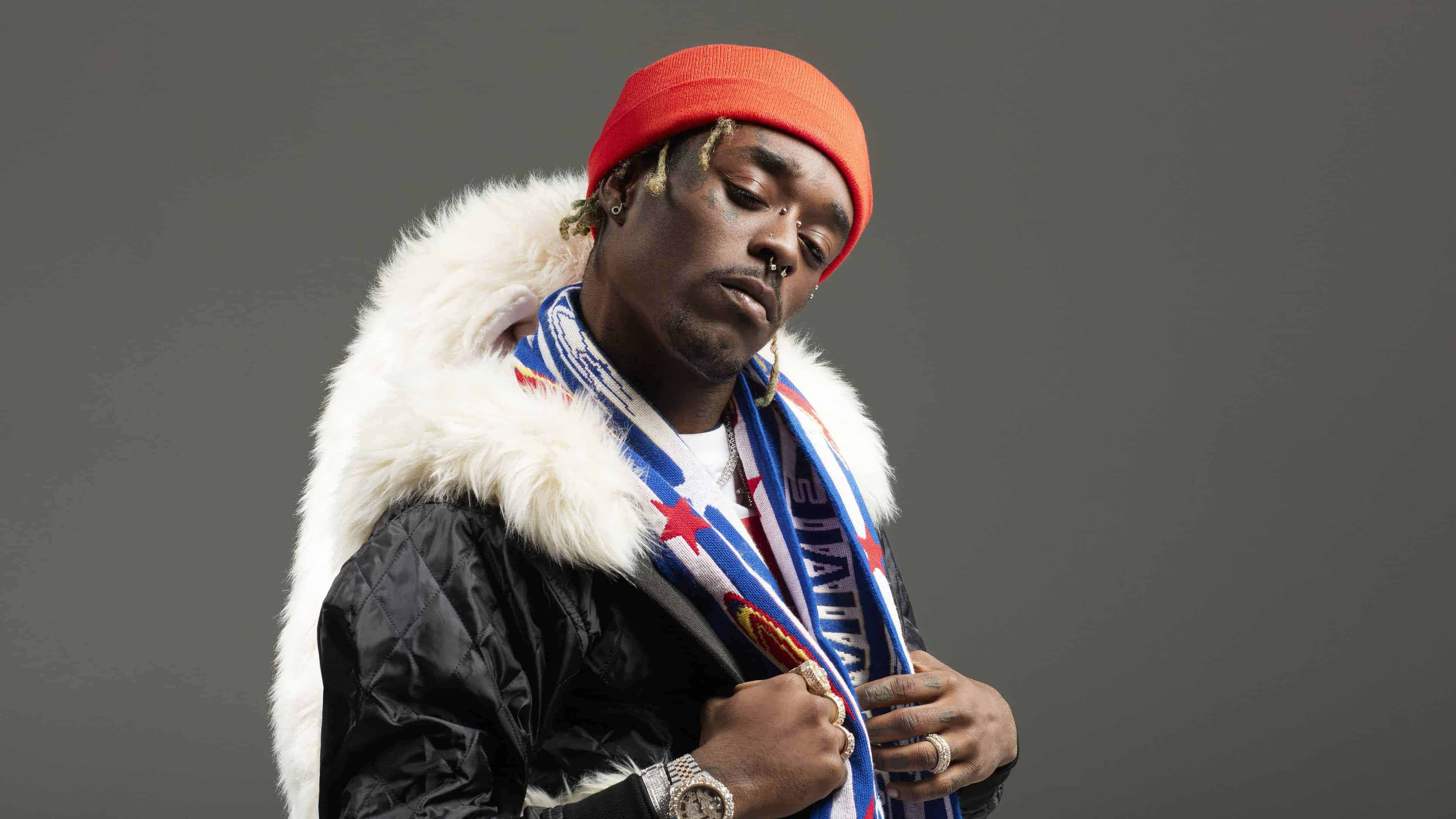 Lil Uzi Vert 4K 桌布 壁紙 — 一個戴紅色毛帽、穿毛領外套的男人，在純灰色背景前向下看。