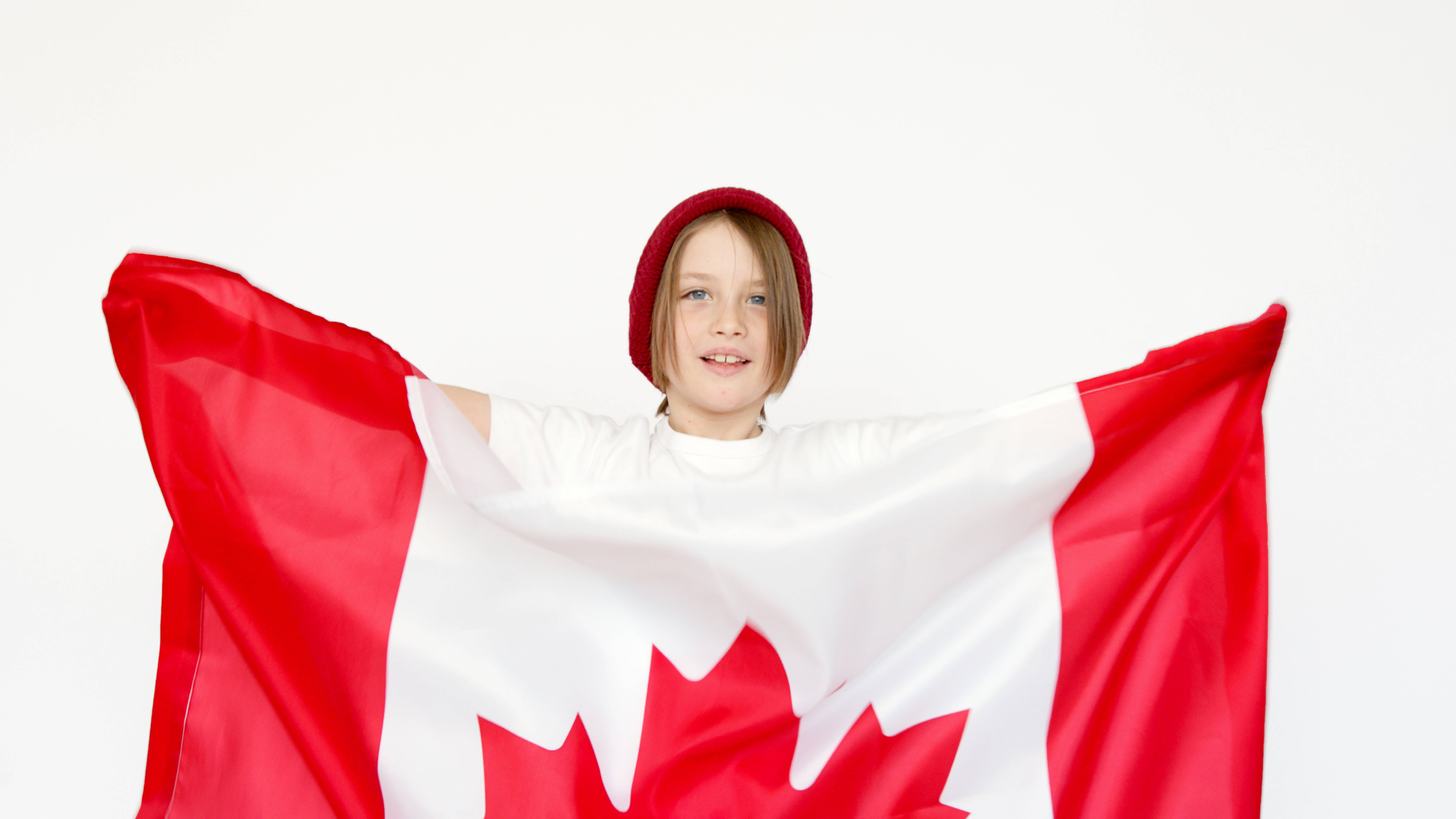 Flag 桌布 壁紙 — 一個戴紅色毛帽的快樂小孩，在純白背景上自豪地拿著加拿大國旗。