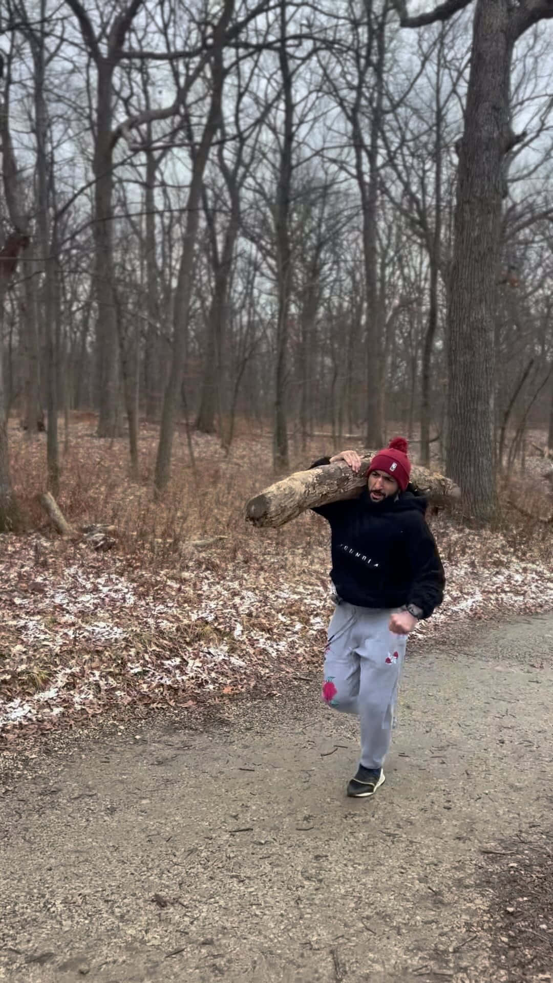 森林 桌布 壁紙 — 一個戴紅色毛帽的男人，在雪地森林小徑上慢跑，肩上扛著一根沉重的木頭。