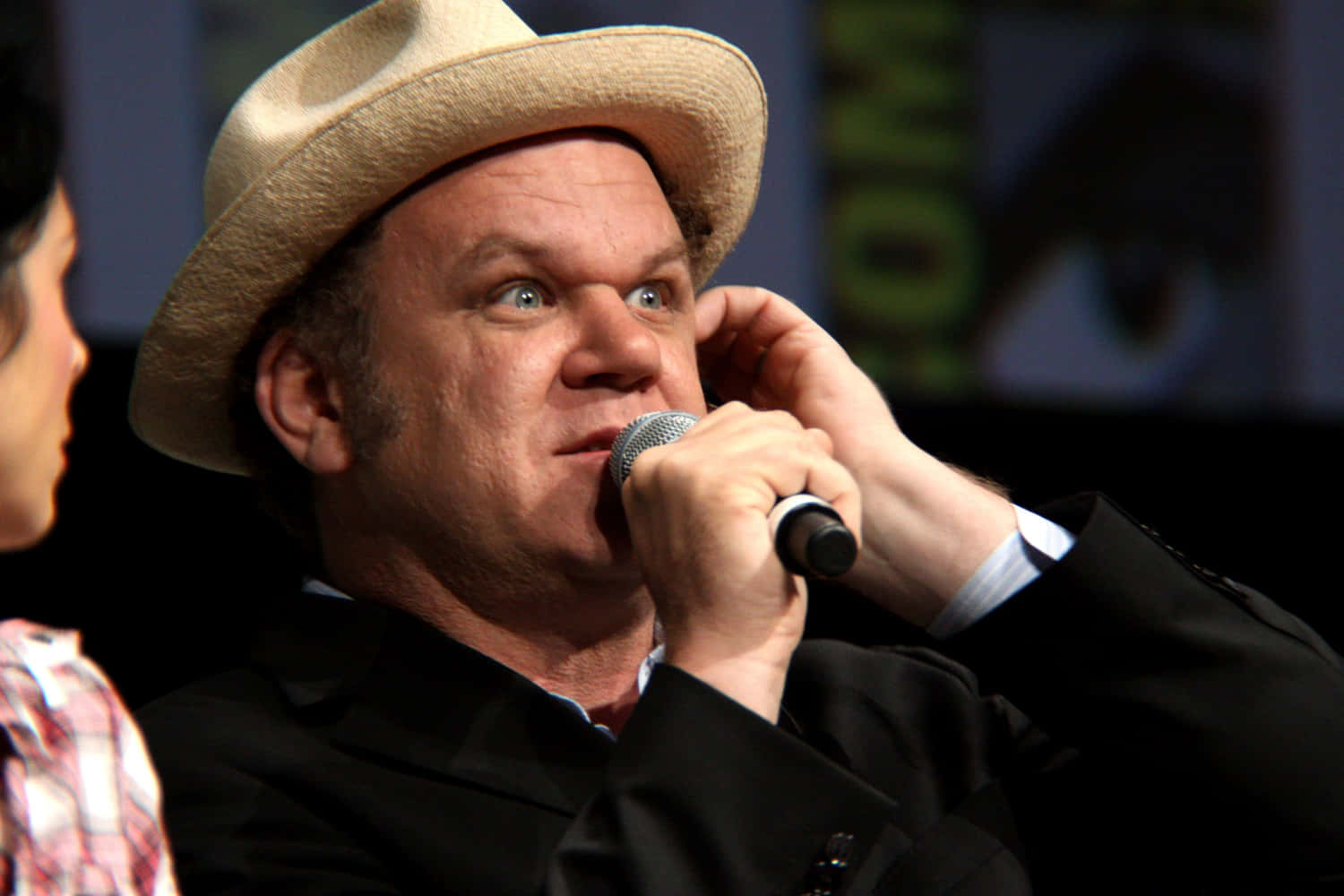 John C. Reilly 桌布 壁紙 — 一個戴草帽的男人在小組討論期間，睜大眼睛、表情驚訝地握著麥克風。