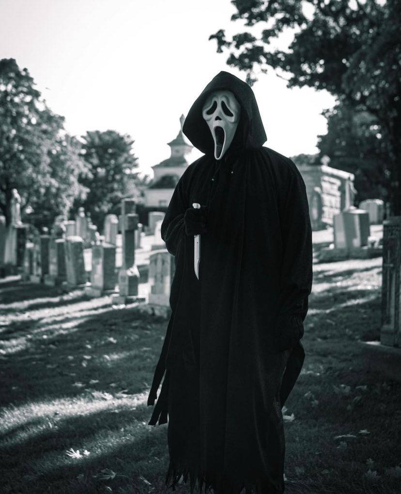 恐怖動圖 桌布 壁紙 — 一個戴著Ghostface面具、身穿黑色長袍的人物，在陽光明媚的墓園中手持刀具，氣勢洶洶地站立。