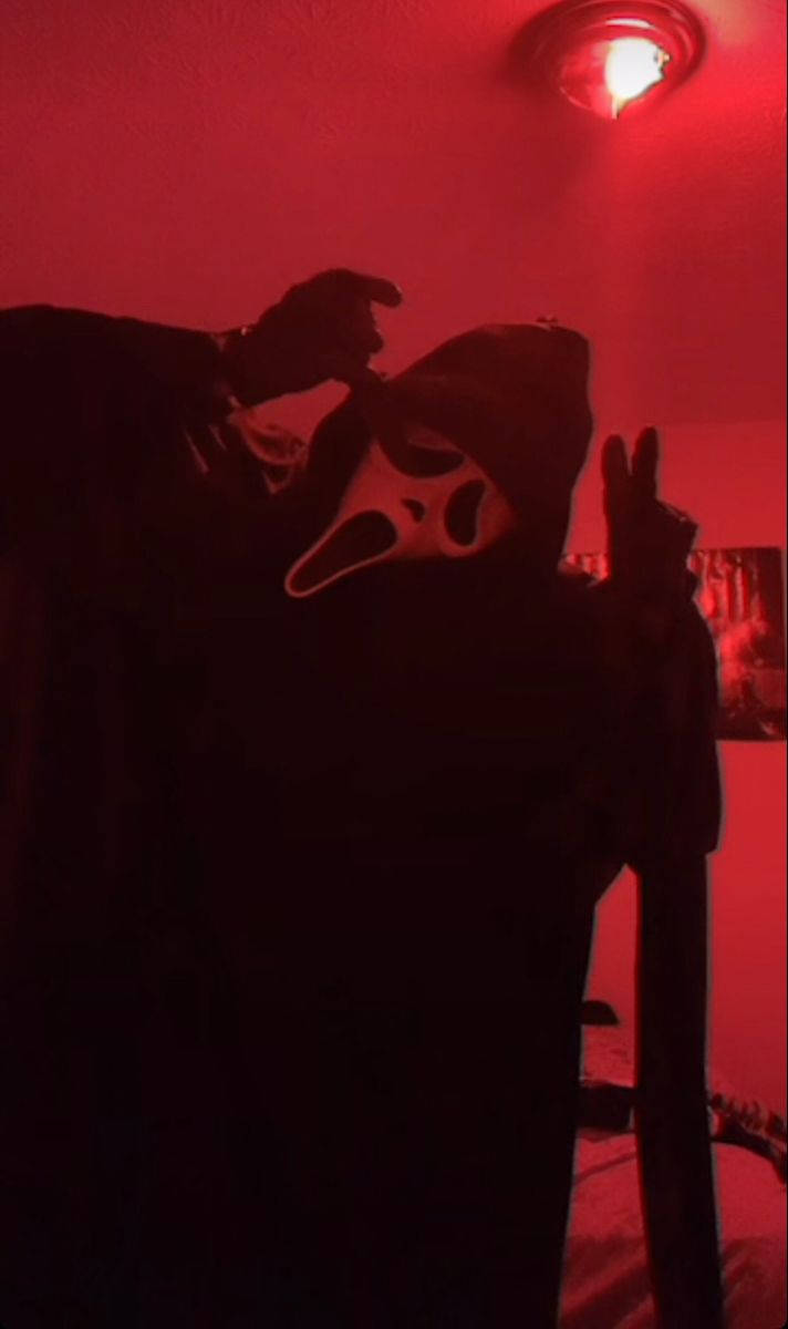 鬼臉殺手 桌布 壁紙 — 一個戴著Ghostface面具的人物站在昏暗的紅色燈光房間中，單手做出和平手勢。