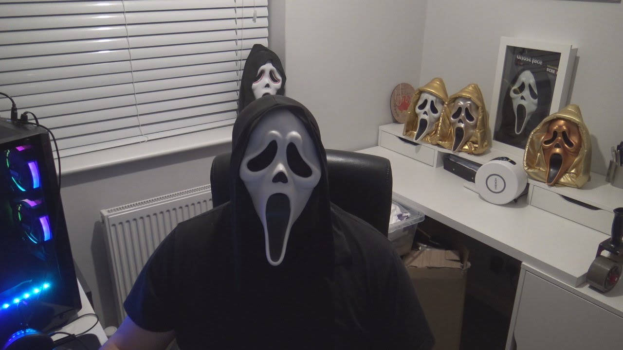 恐怖動圖 桌布 壁紙 — 一個戴著Ghostface面具的人坐在一張被恐怖電影紀念品和發光電腦設備包圍的書桌前。