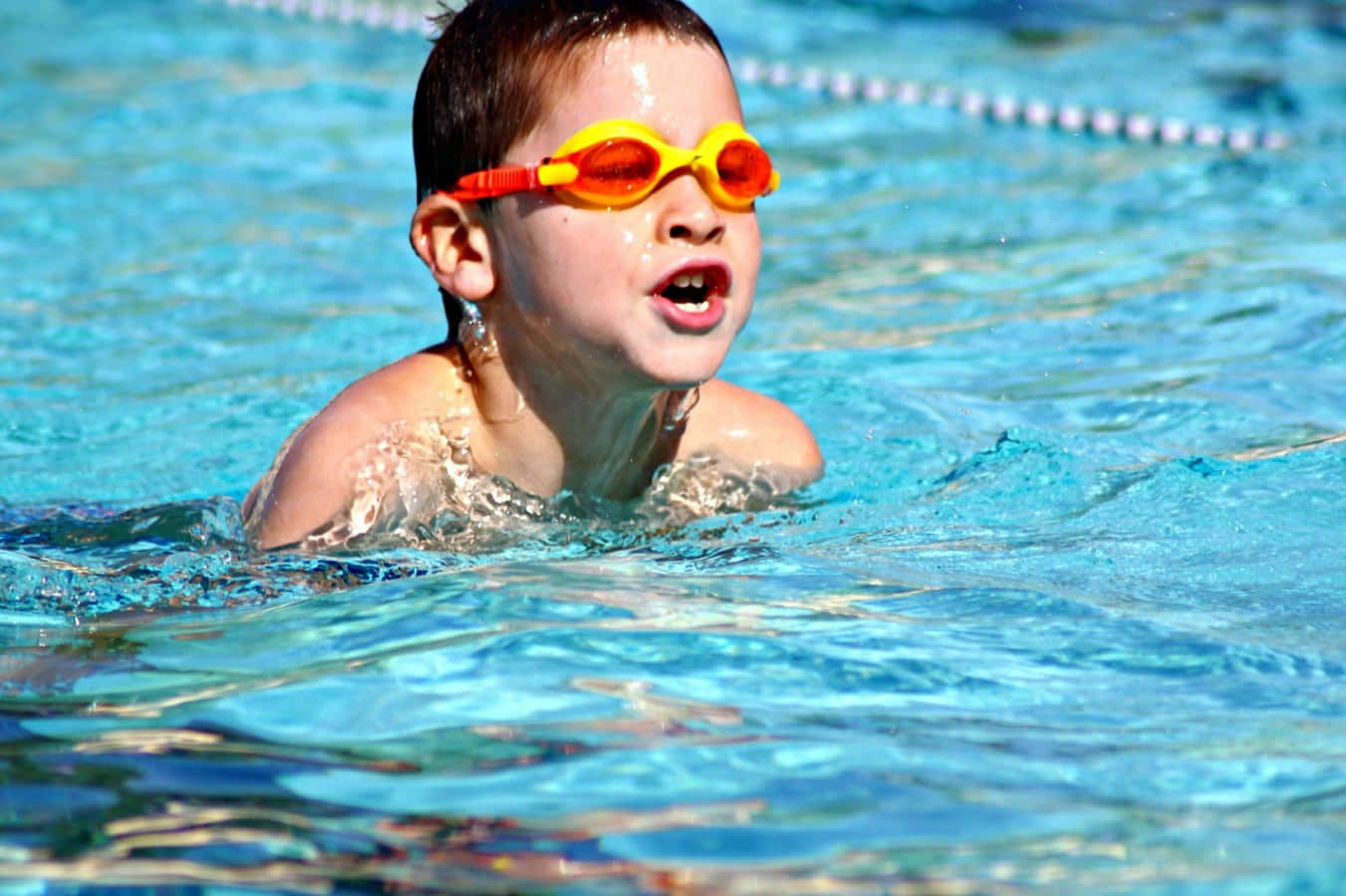 Swimming 桌布 壁紙 — 一個戴著亮橙色泳鏡的小男孩在晴朗的天空下，在清澈的藍色泳池水中快樂地潑水。