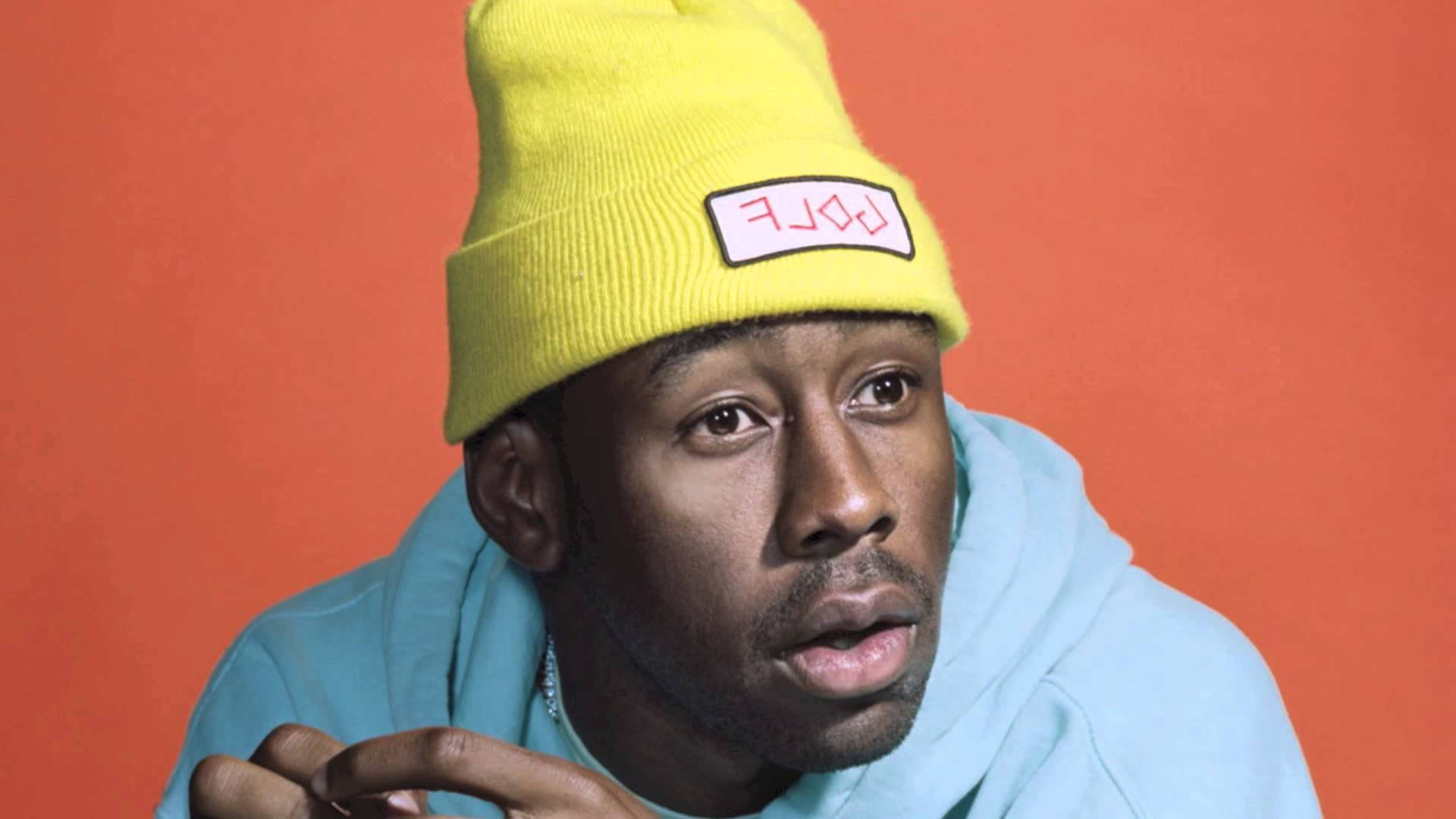 tyler 桌布 壁紙 — 一個戴著亮黃色毛帽和淺藍色連帽衫的男人在橙色背景前擺姿勢，表情若有所思。