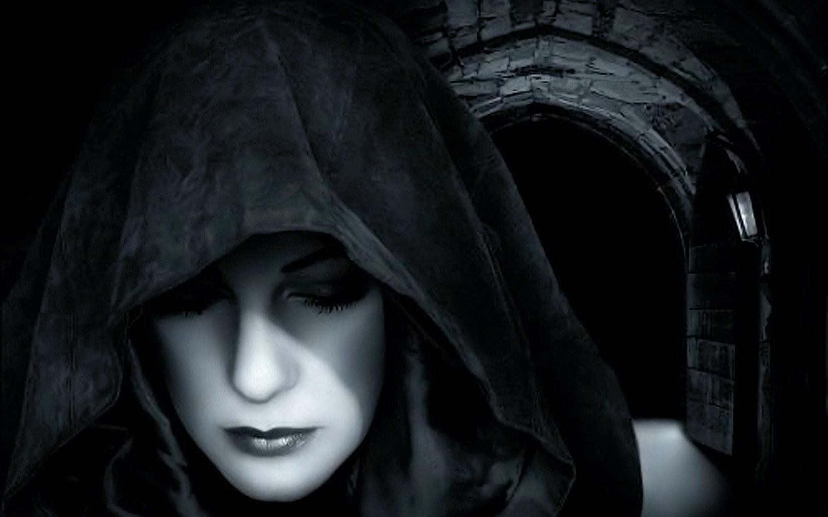 Dark Gothic 桌布 壁紙 — 一個戴著兜帽、皮膚蒼白、眼睛漆黑的身影在昏暗的石拱門中向下凝視，被一束光照亮。