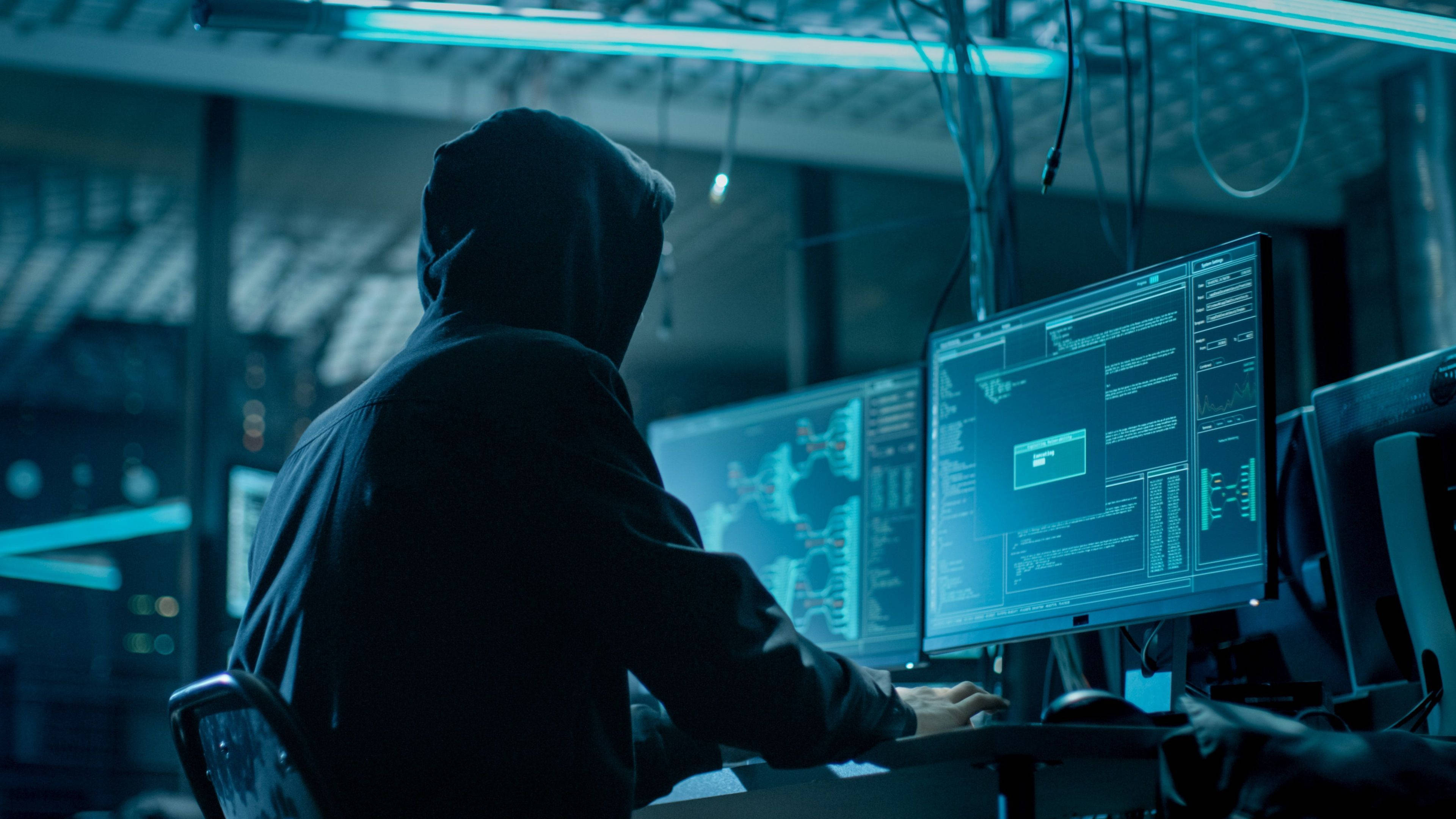 Cyber 桌布 壁紙 — 一個戴著兜帽的人坐在昏暗的伺服器機房中，沐浴在藍光下，在多個顯示複雜數據的電腦顯示器上專注工作。