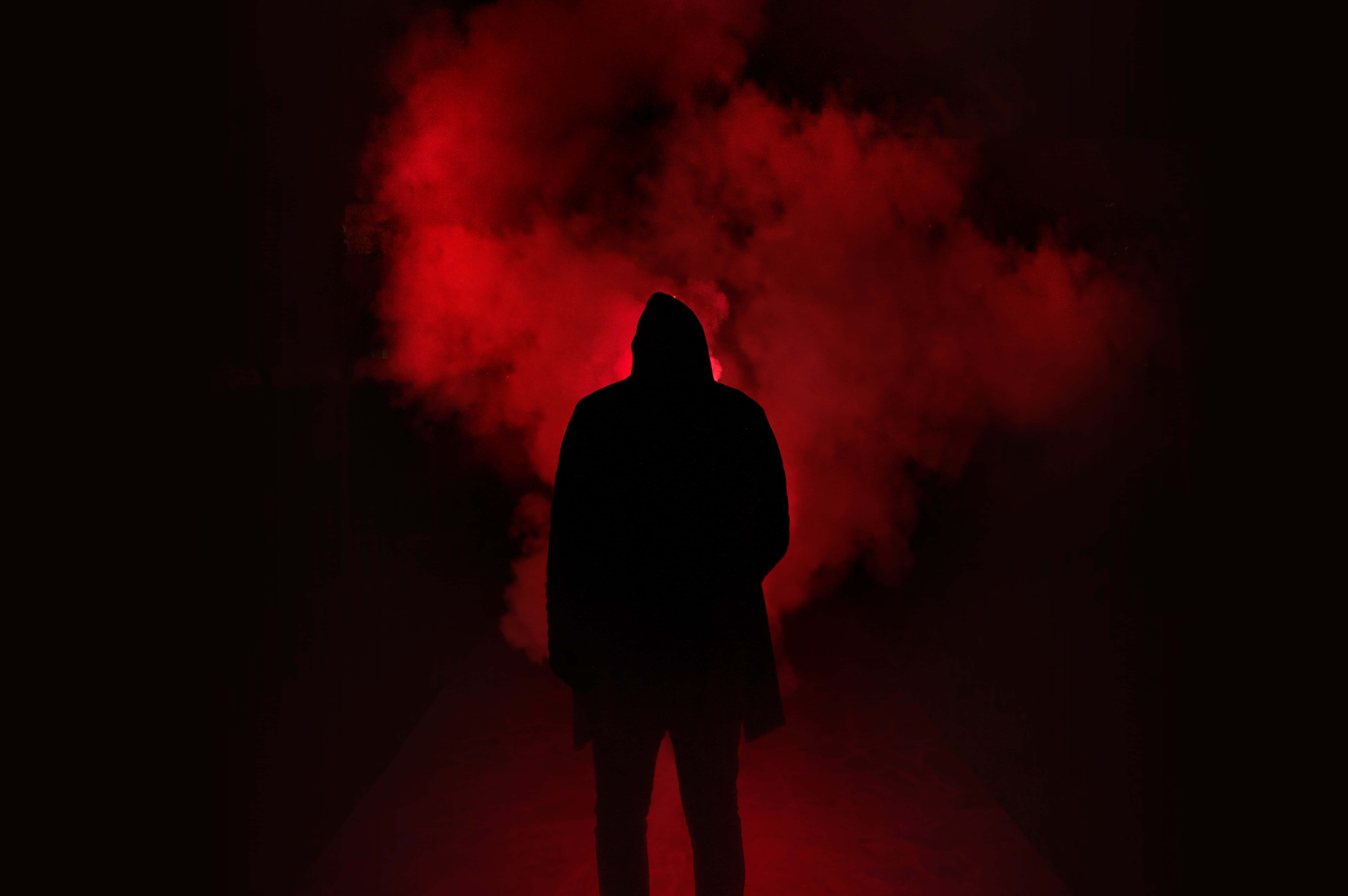 Black And Red 桌布 壁紙 — 一個戴著兜帽的人影站在黑暗的走廊中，被旋轉的紅煙照亮。