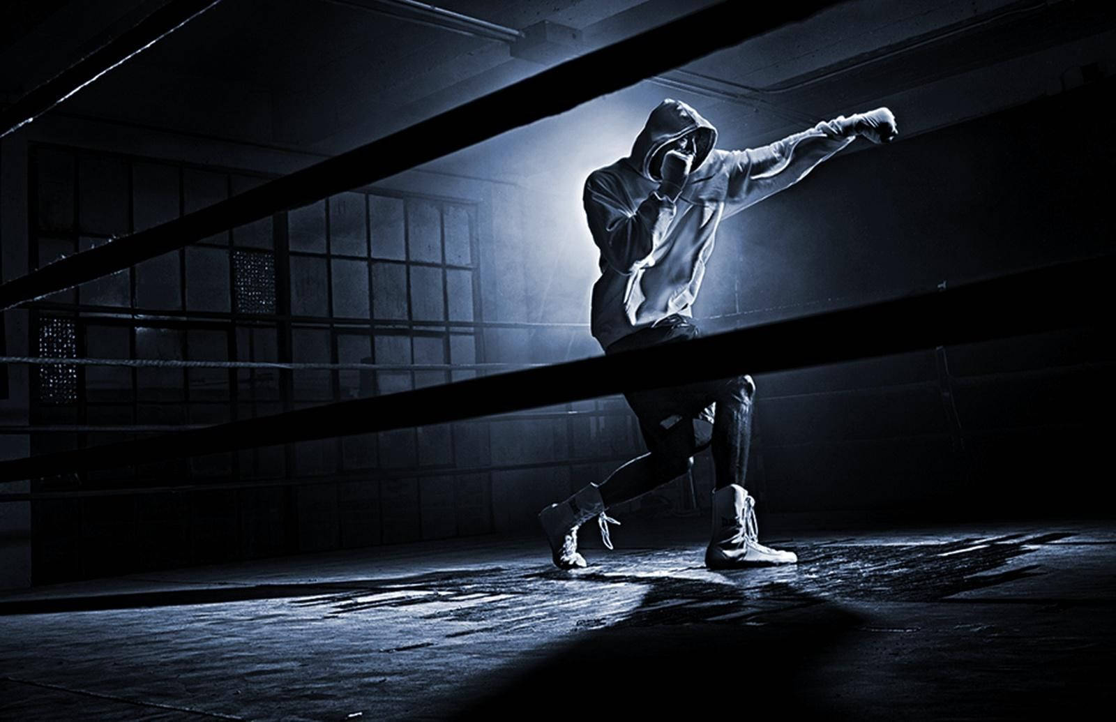 Boxing 桌布 壁紙 — 一個戴著兜帽的人物以拳擊姿勢站在昏暗骯髒的擂台上，被單一盞聚光燈照亮。
