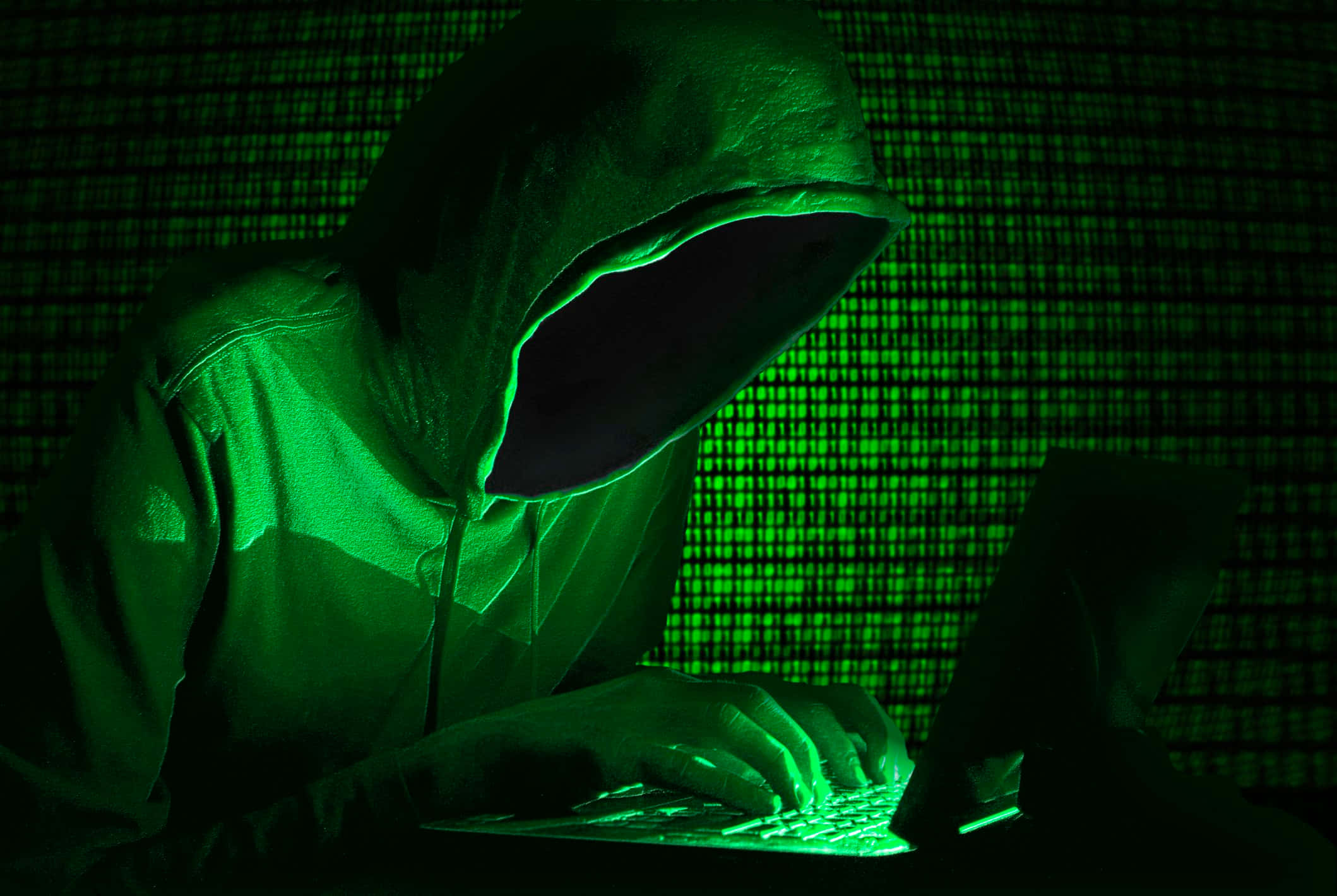 Dark Web 桌布 壁紙 — 一個戴著兜帽的人物在詭異的綠色光芒下，於數位網格背景前打字。