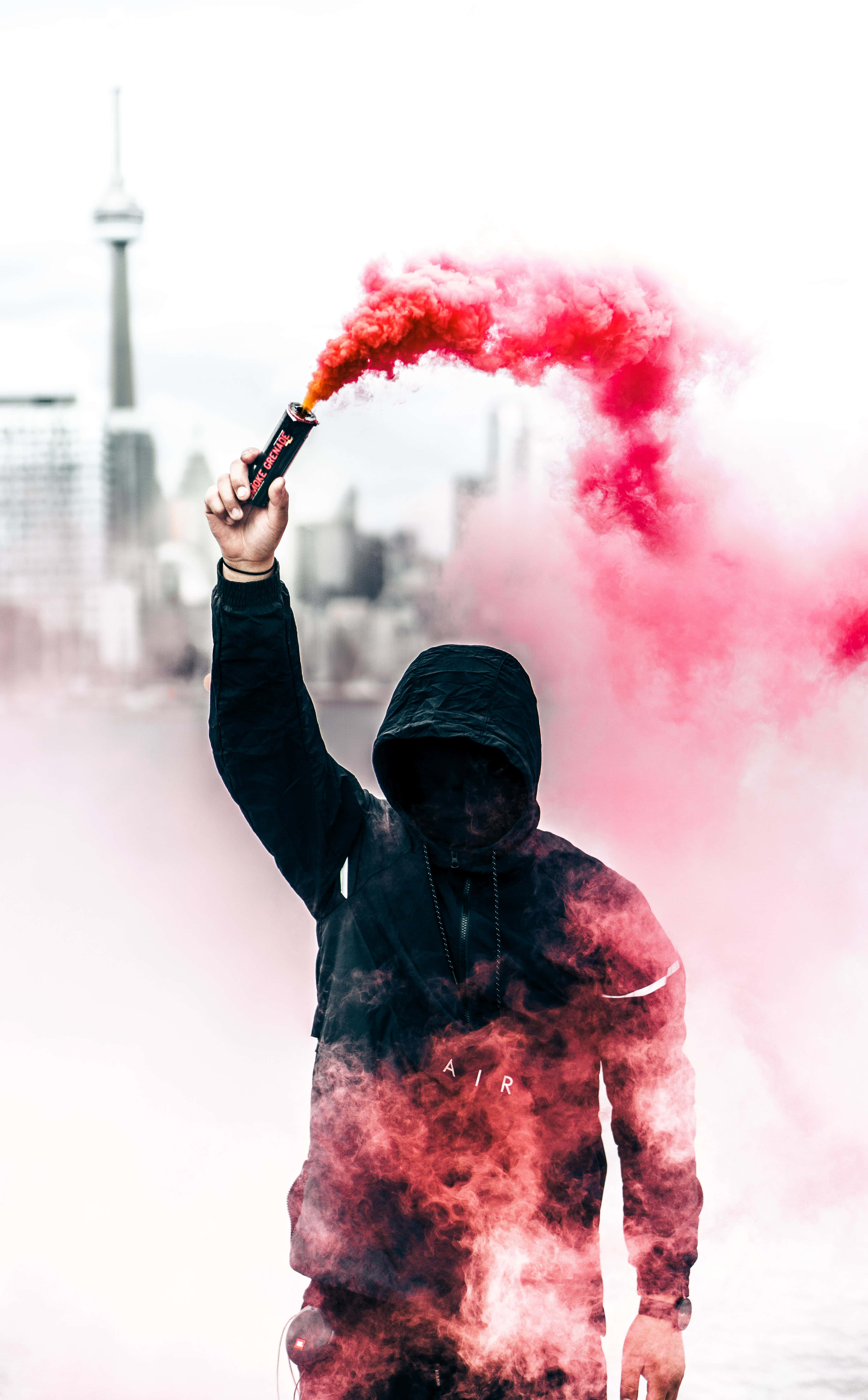 Flare 桌布 壁紙 — 一個戴著兜帽的人物手持紅色煙霧信號彈，背景是模糊的城市天際線，遠處可見一座塔。