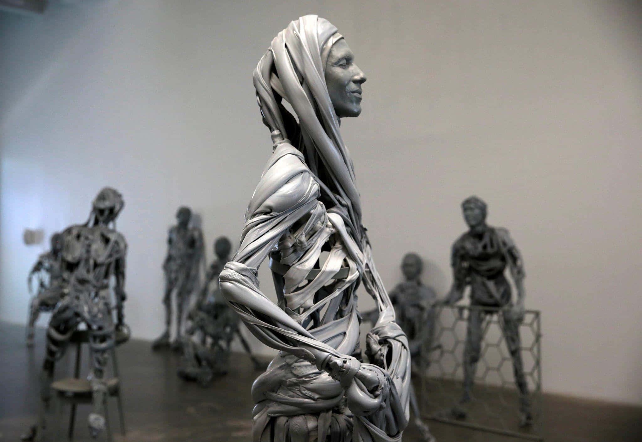 Kinetic Art 桌布 壁紙 — 一個戴著兜帽的人物銀色雕塑，在極簡主義畫廊空間中，側面站立於其他金屬人形之間。
