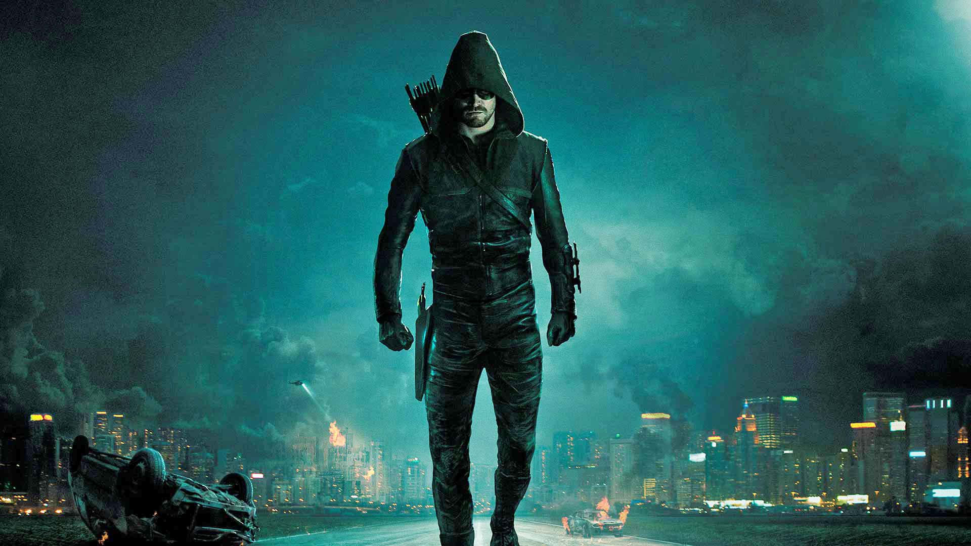 Green Arrow 桌布 壁紙 — 一個戴著兜帽的義警站在夜晚被雨水浸濕的城市街道上，附近有一輛翻倒的汽車，身後是燃燒的天際線。