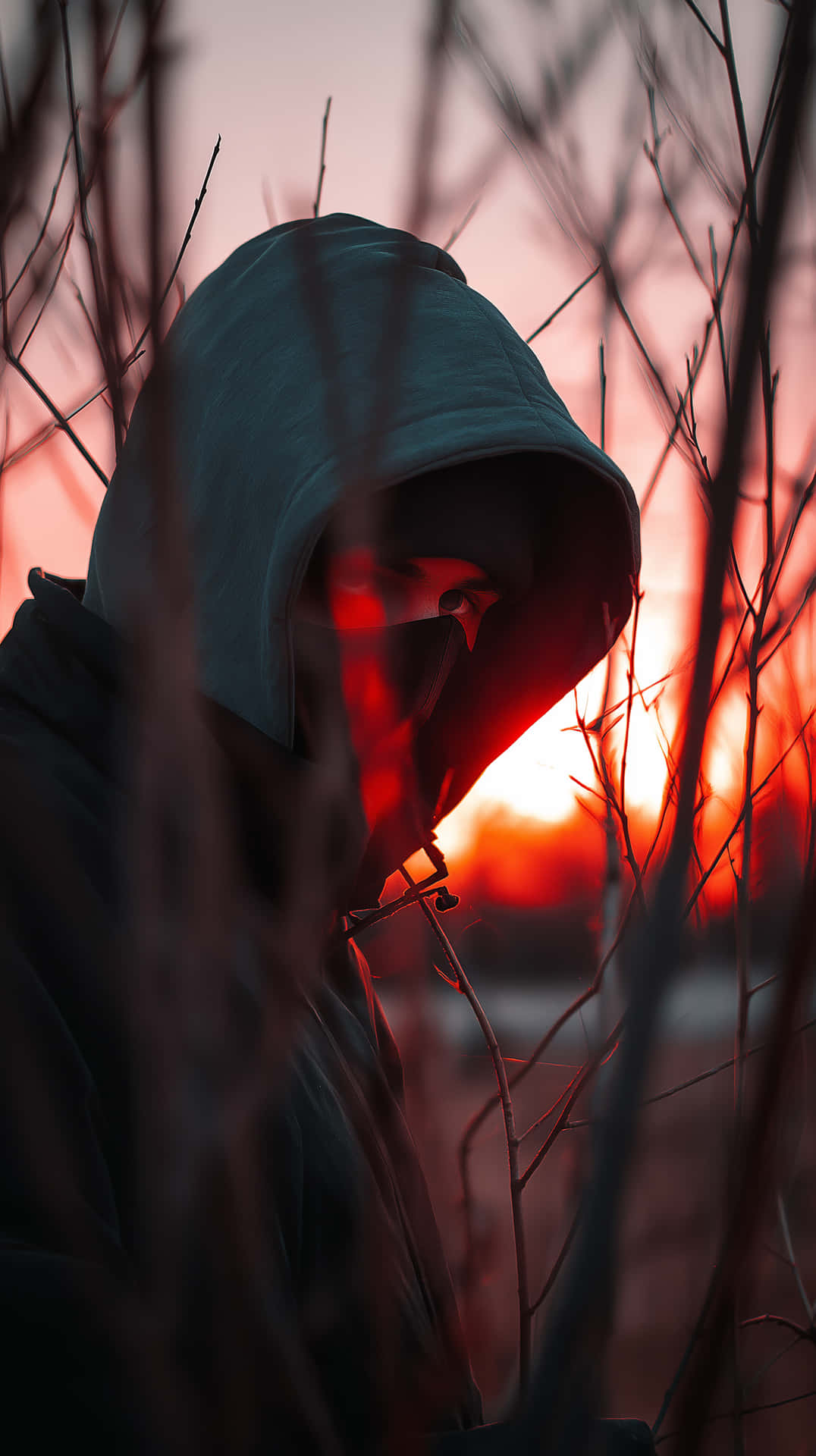 Hooded Silhouette at Sunset - Enigmatic Figure Among Branches 壁紙 — 一個戴著兜帽的身影，雙眼發出紅光，透過光禿的樹枝凝視著熾熱的日落。