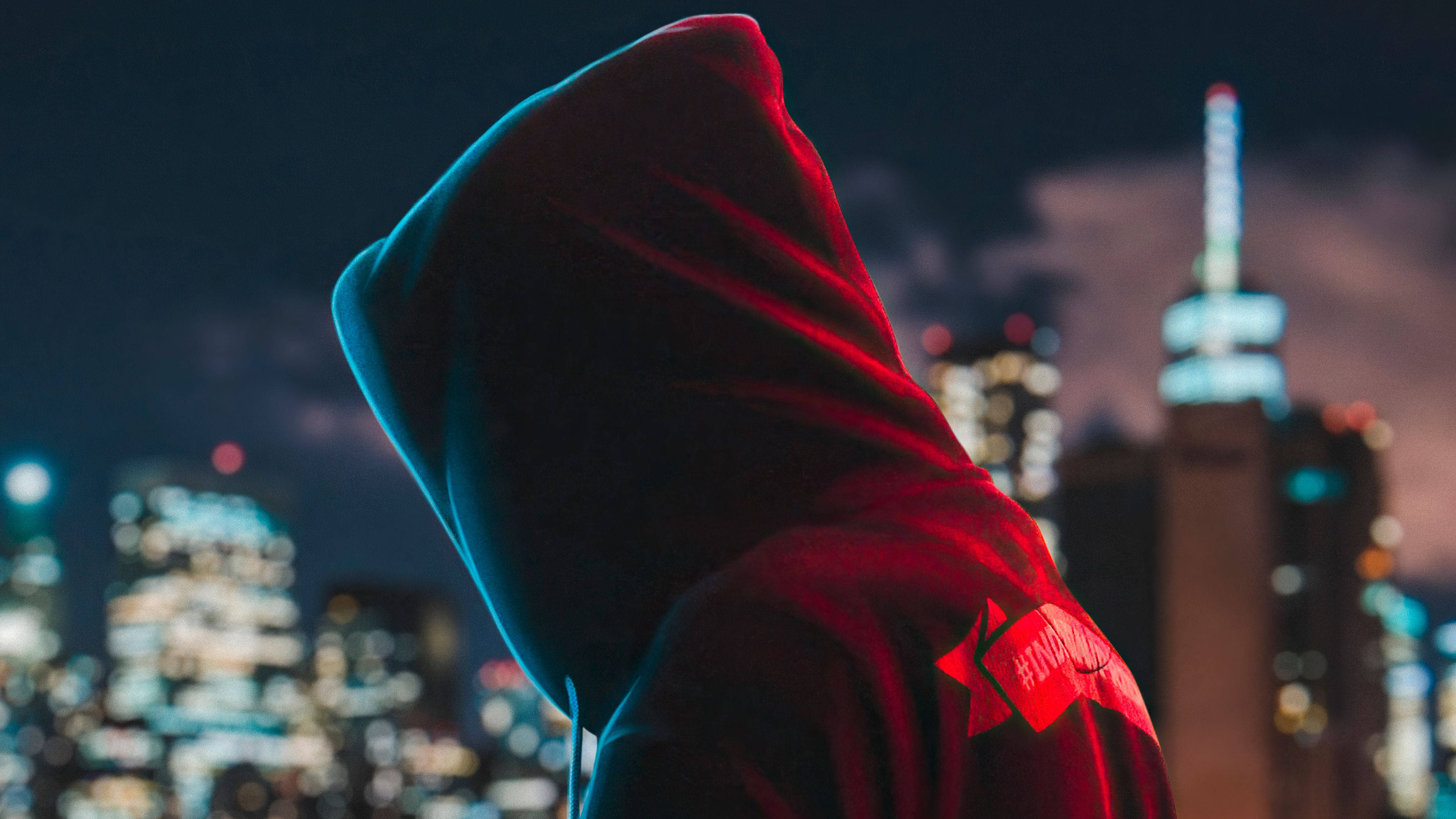 Hoodie 桌布 壁紙 — 一個戴著兜帽的身影在夜晚以霓虹燈照亮的天際線為背景，呈現剪影。