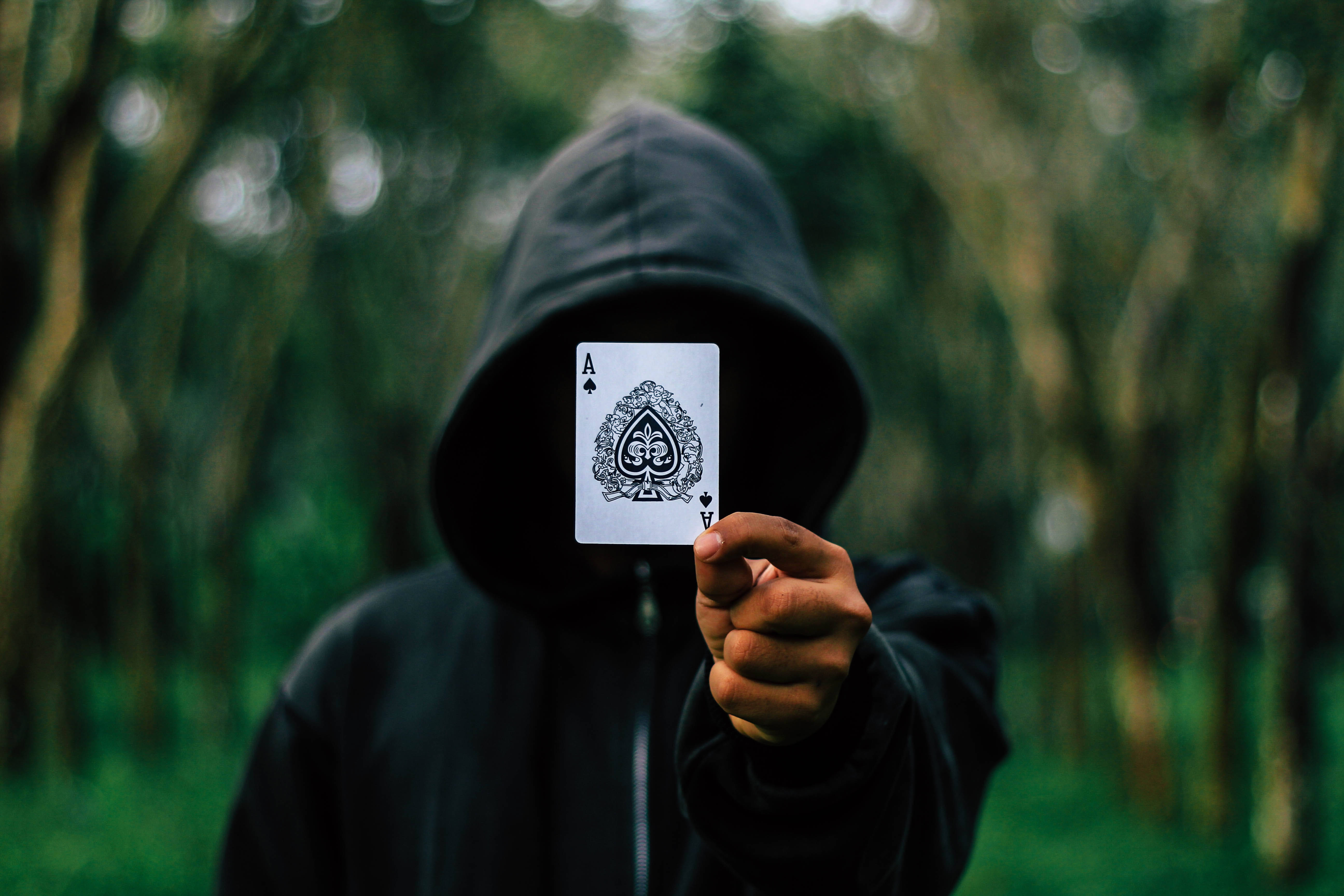 Poker 桌布 壁紙 — 一個戴著兜帽的身影在森林中舉起一張黑桃A牌遮住臉，做出神秘的姿態。