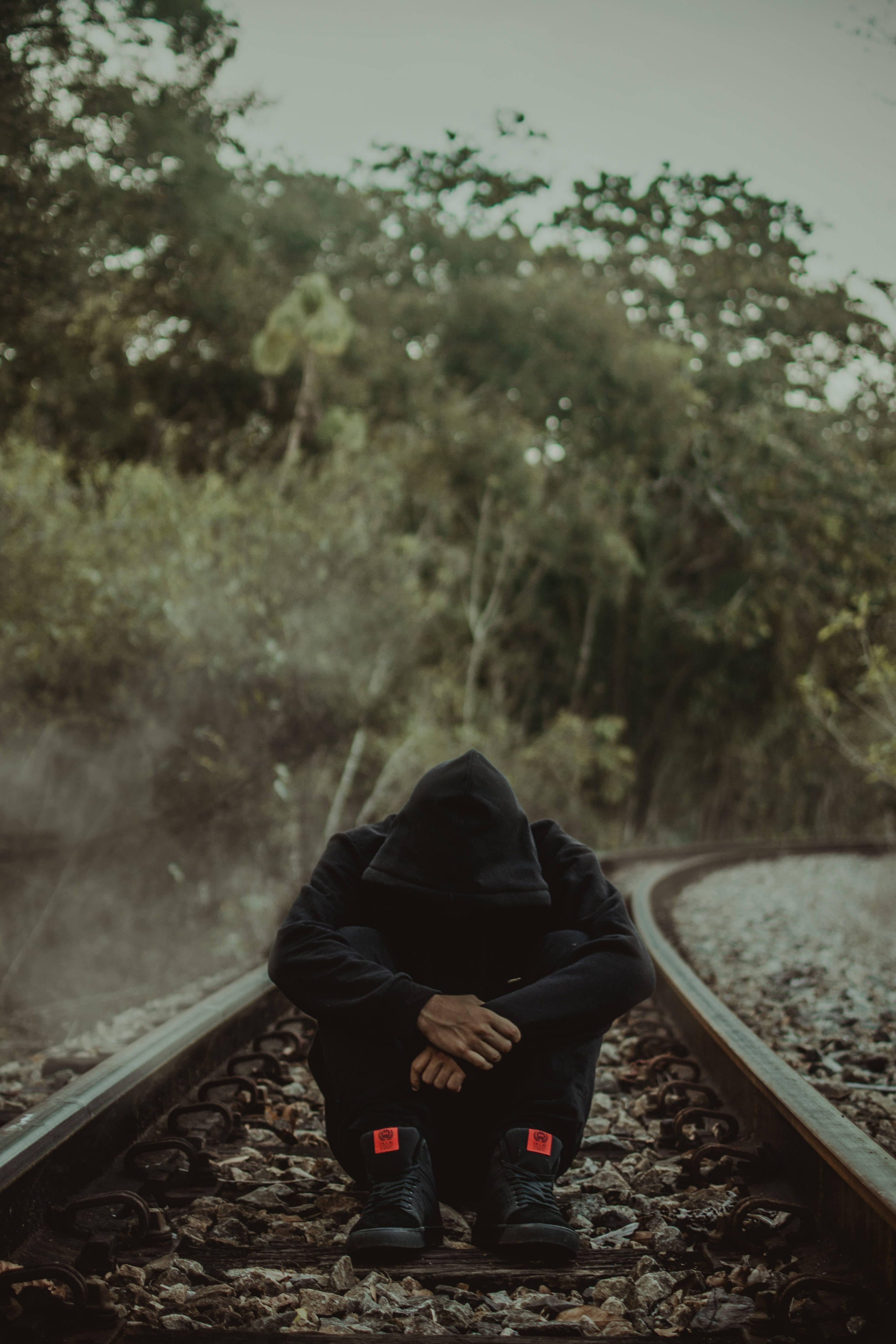 Alone 桌布 壁紙 — 一個戴著兜帽的身影獨自坐在鐵軌上，周圍是霧氣瀰漫的綠色樹葉，天空多雲。
