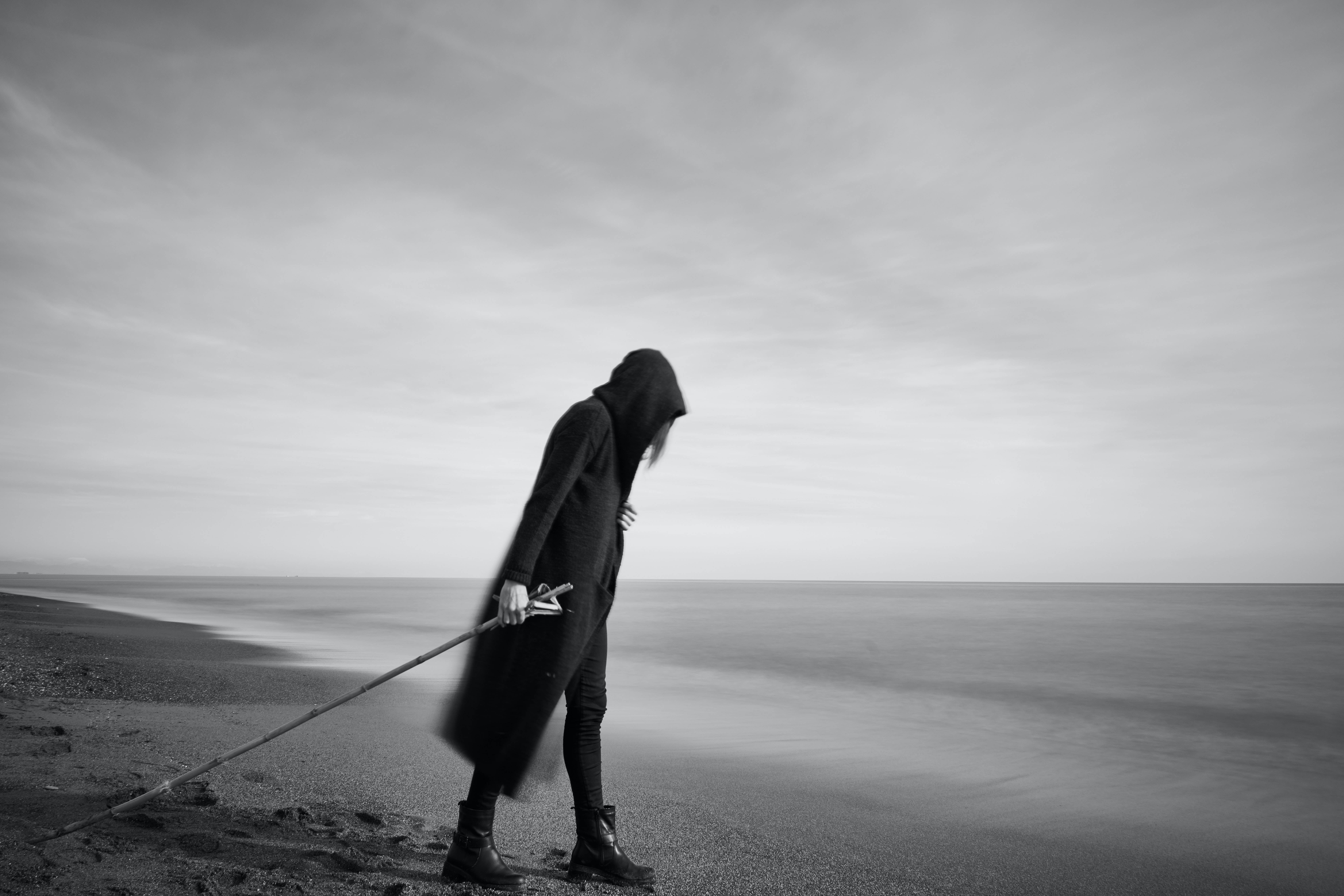 海灘 桌布 壁紙 — 一個戴著兜帽的身影獨自站在霧氣瀰漫的海灘上，手持長杖，背景中的海浪模糊不清。