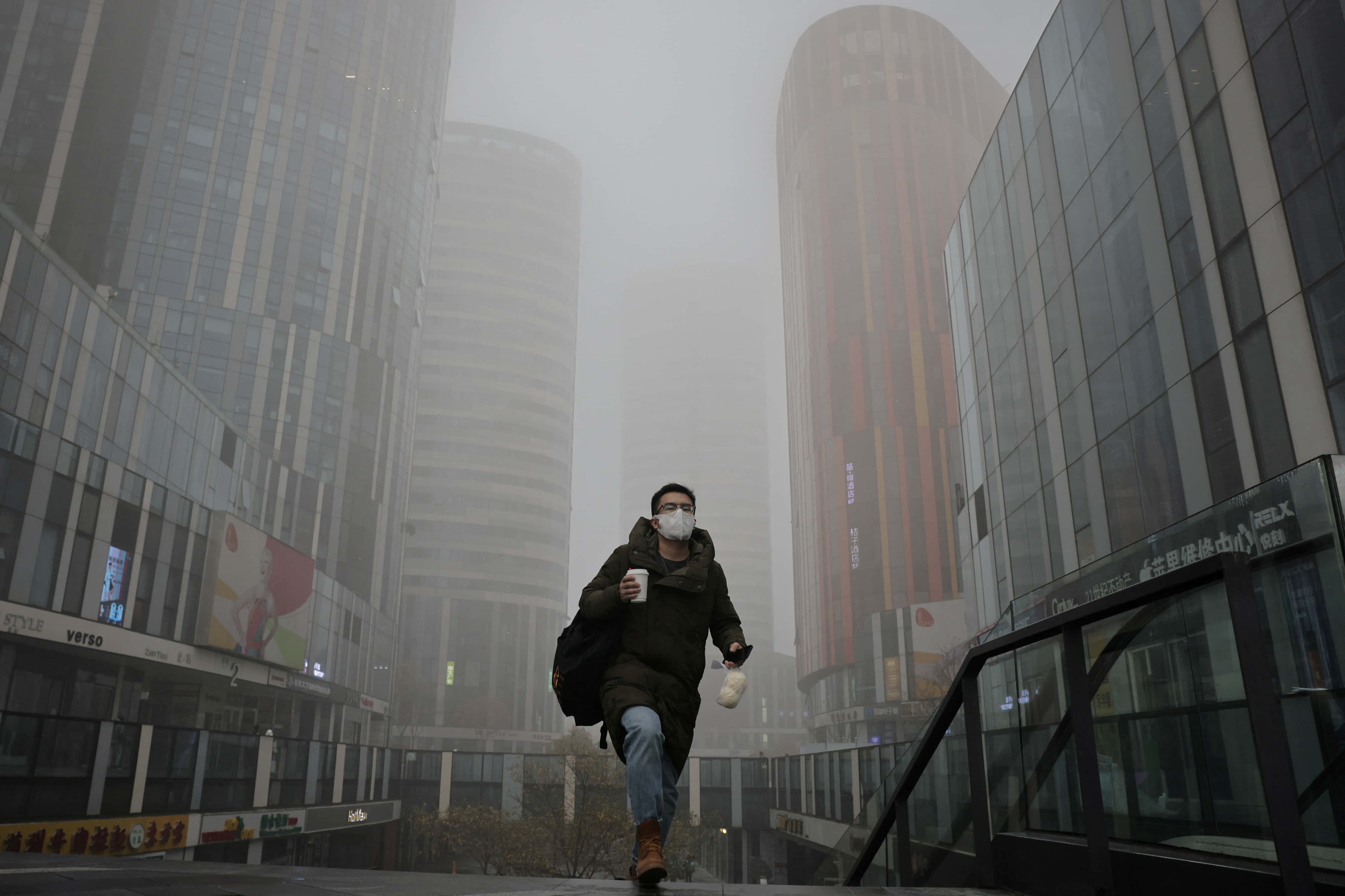 中國 桌布 壁紙 — 一個戴著口罩的人匆匆穿過霧氣瀰漫的都市峽谷，兩旁是高聳的玻璃摩天大樓。