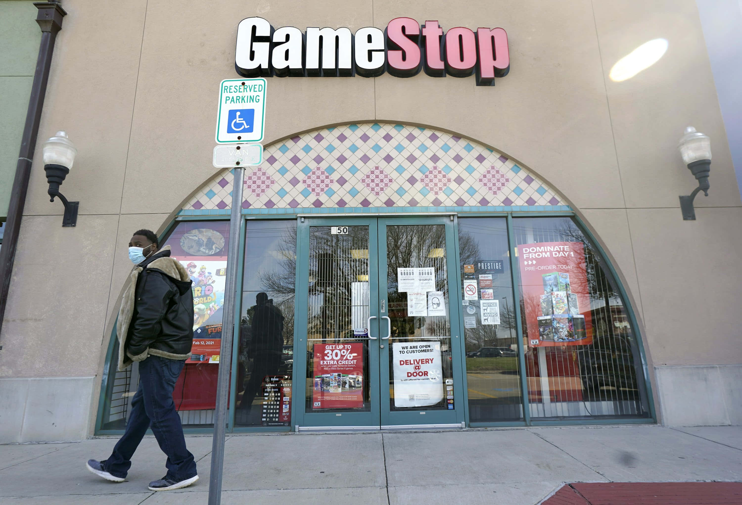 GameStop 桌布 壁紙 — 一個戴著口罩的人走過一間有玻璃門和宣傳海報的GameStop商店，背景是明亮的白天天空。