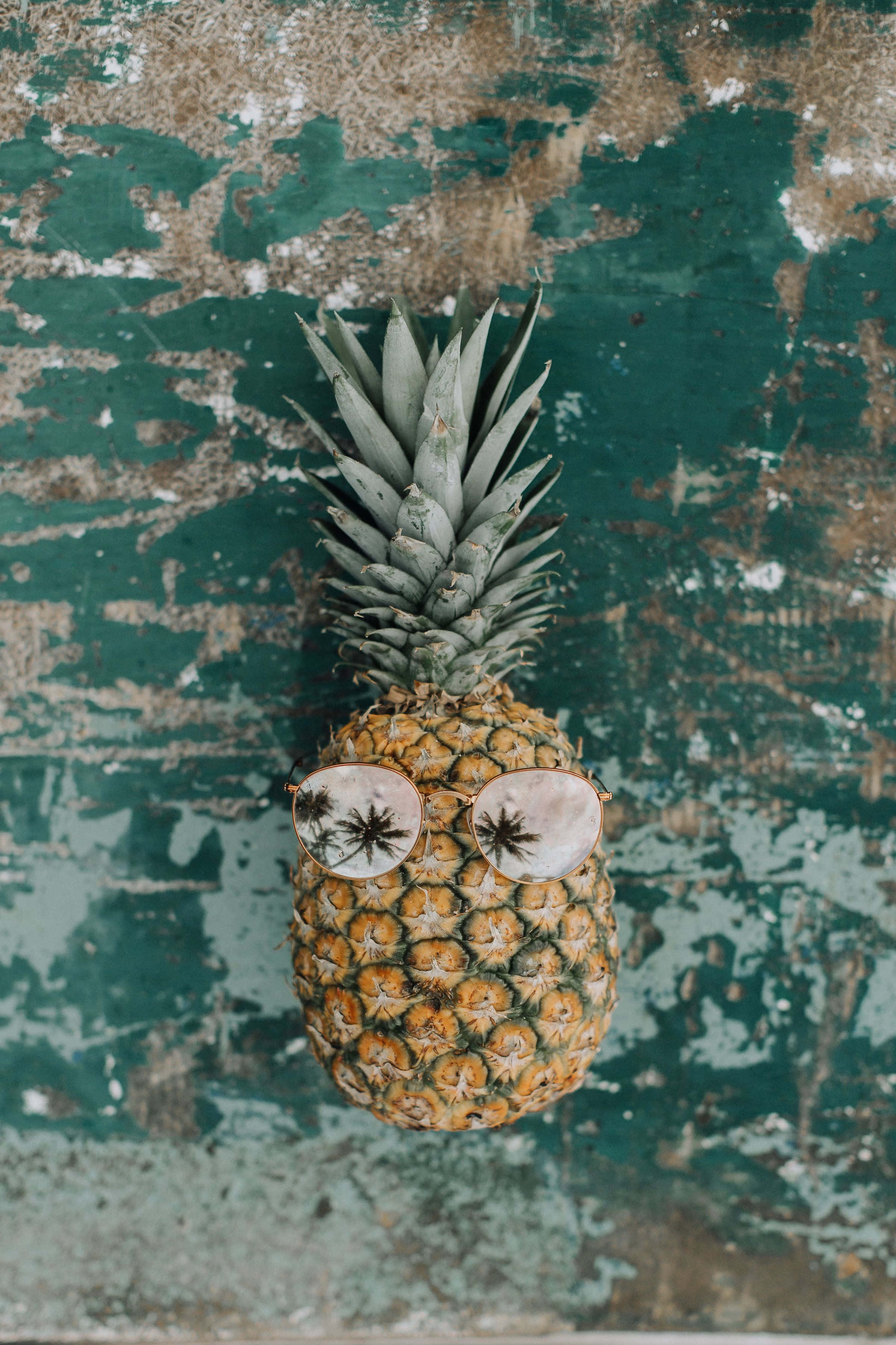 Pineapple 桌布 壁紙 — 一個戴著圓形太陽眼鏡的鳳梨靠在一幅風化的青綠色和棕色剝落牆上。