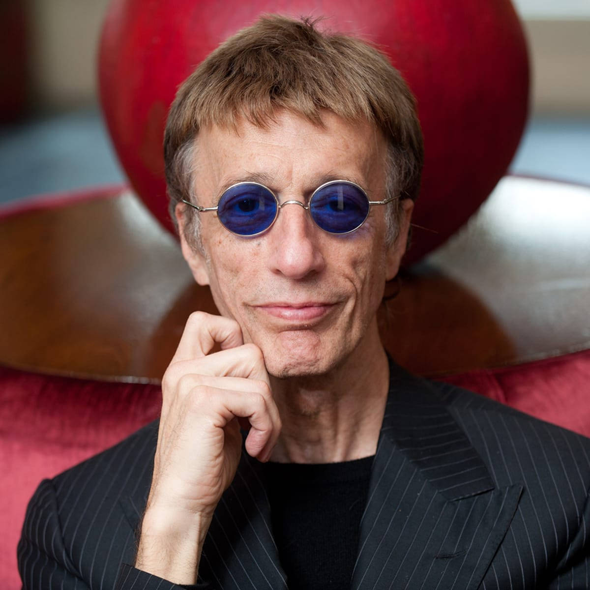 Celebrity 桌布 壁紙 — 一個戴著圓形藍色太陽眼鏡和細條紋西裝的男人，將下巴擱在手上，靠著一個紅色球形雕塑。