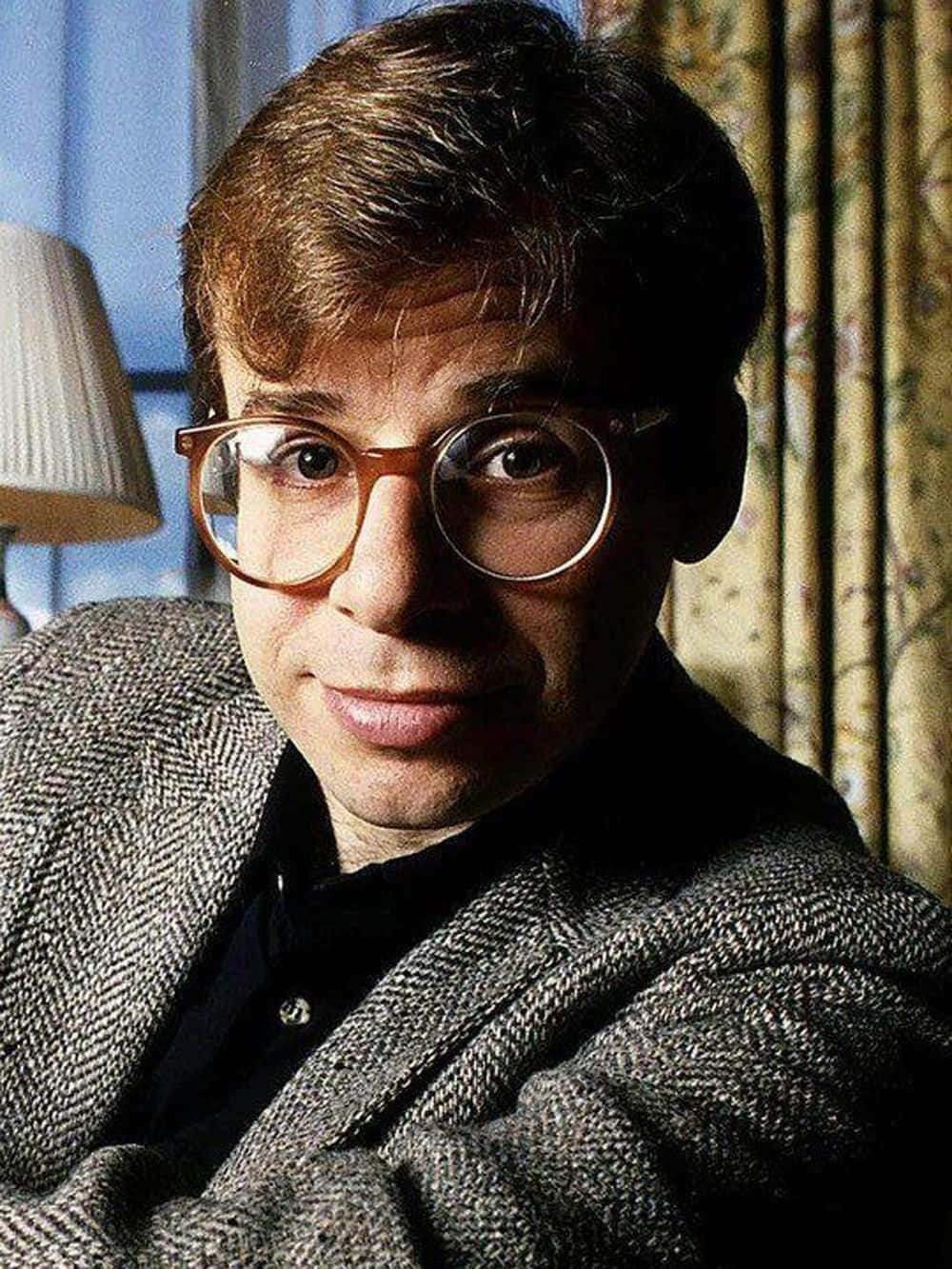rick moranis 桌布 壁紙 — 一個戴著圓框眼鏡和粗花呢外套的男人在溫暖燈光的室內環境中直視鏡頭。