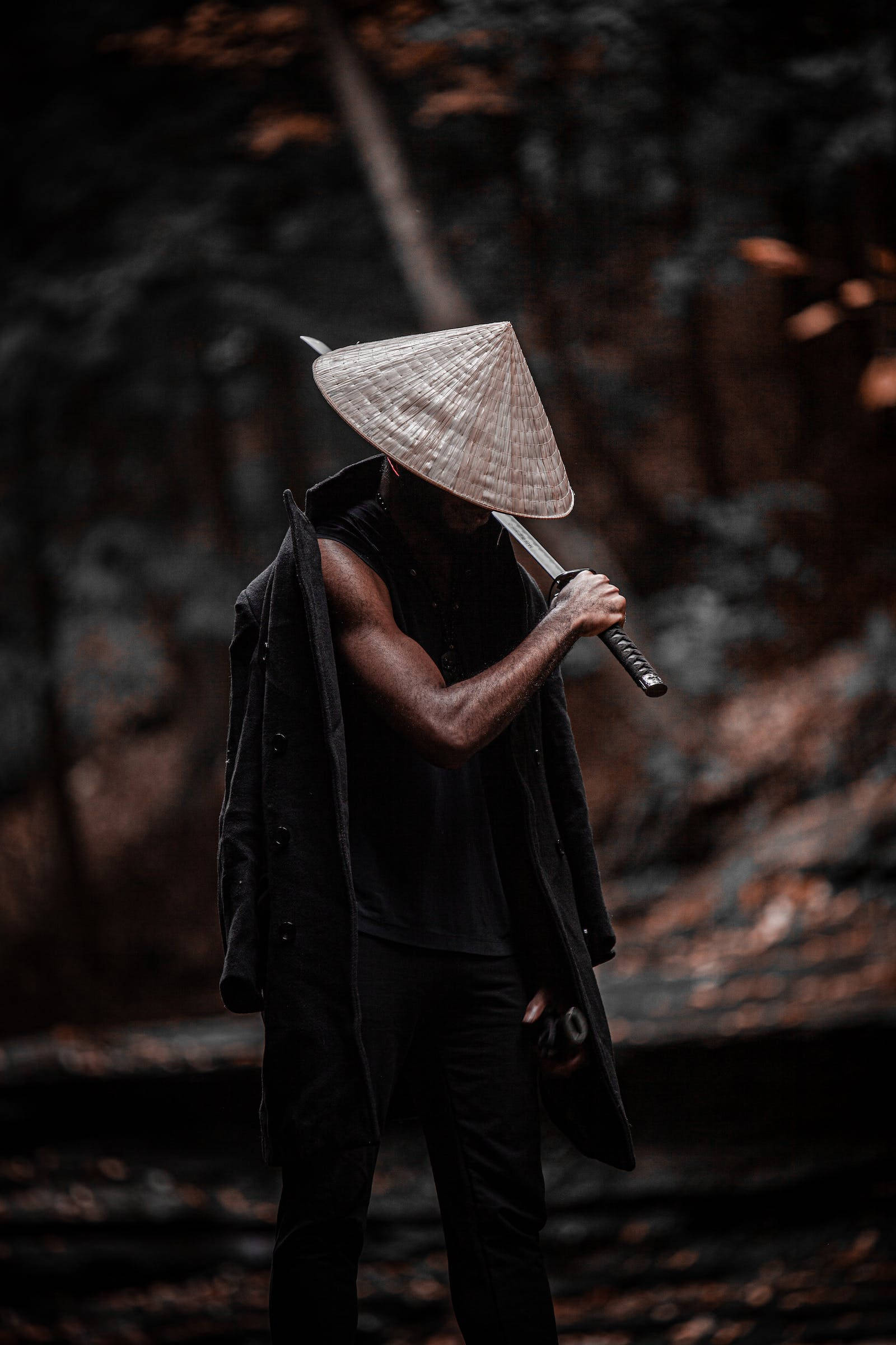 桌布 壁紙 — 一個戴著圓錐形帽子、身穿黑色外套的人物，在昏暗的森林中手持劍，周圍有秋葉。