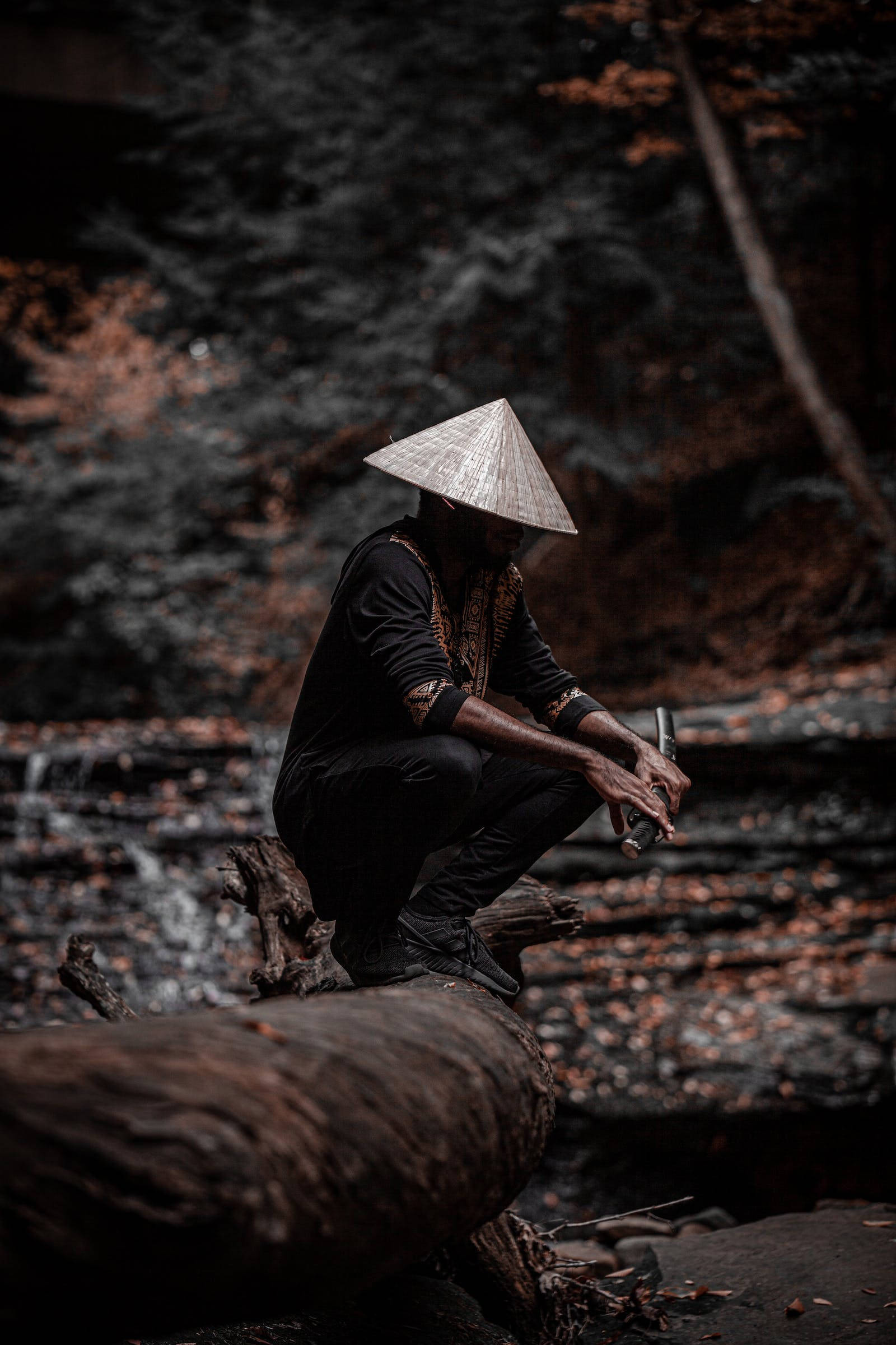 桌布 壁紙 — 一個戴著圓錐形帽子的人蹲在黑暗森林中一條長滿苔蘚的小溪旁邊的倒下的木頭上。