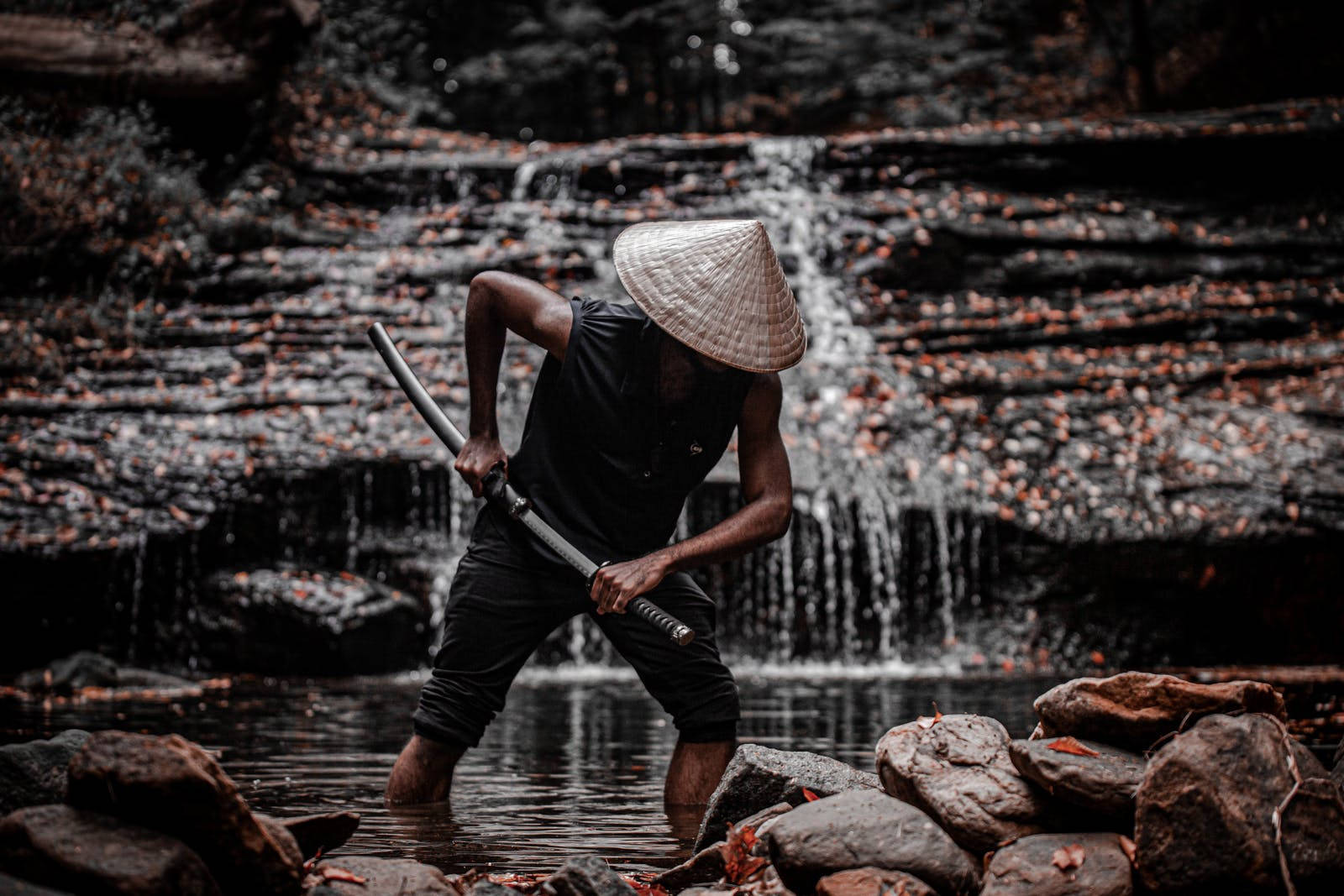桌布 壁紙 — 一個戴著圓錐形帽子的男人在淺水中揮舞著武士刀，面前是瀑布，周圍環繞著秋葉。