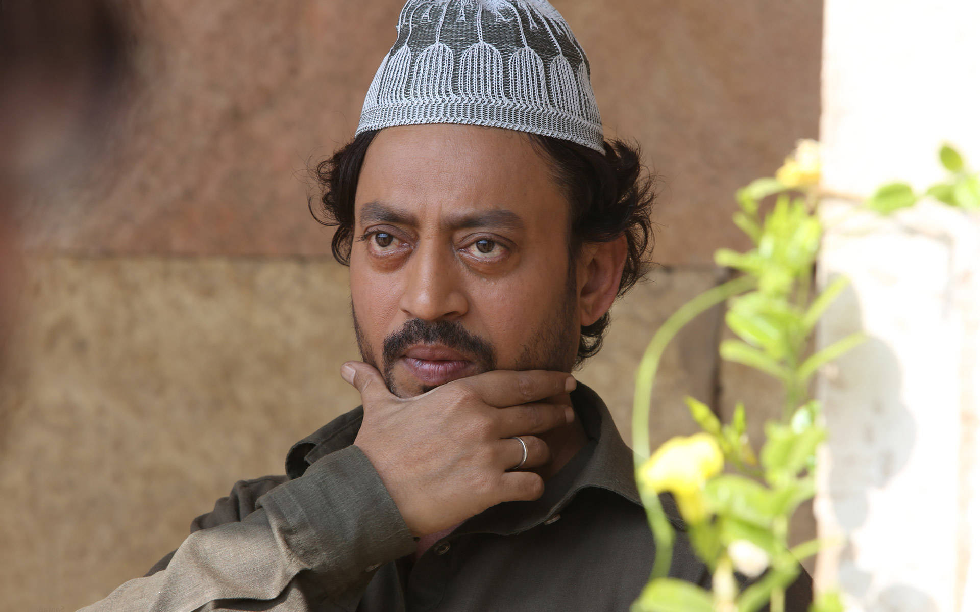 Irrfan Khan 桌布 壁紙 — 一個戴著圖案帽的男人，手托下巴，若有所思地凝視著旁邊一株開花的植物。